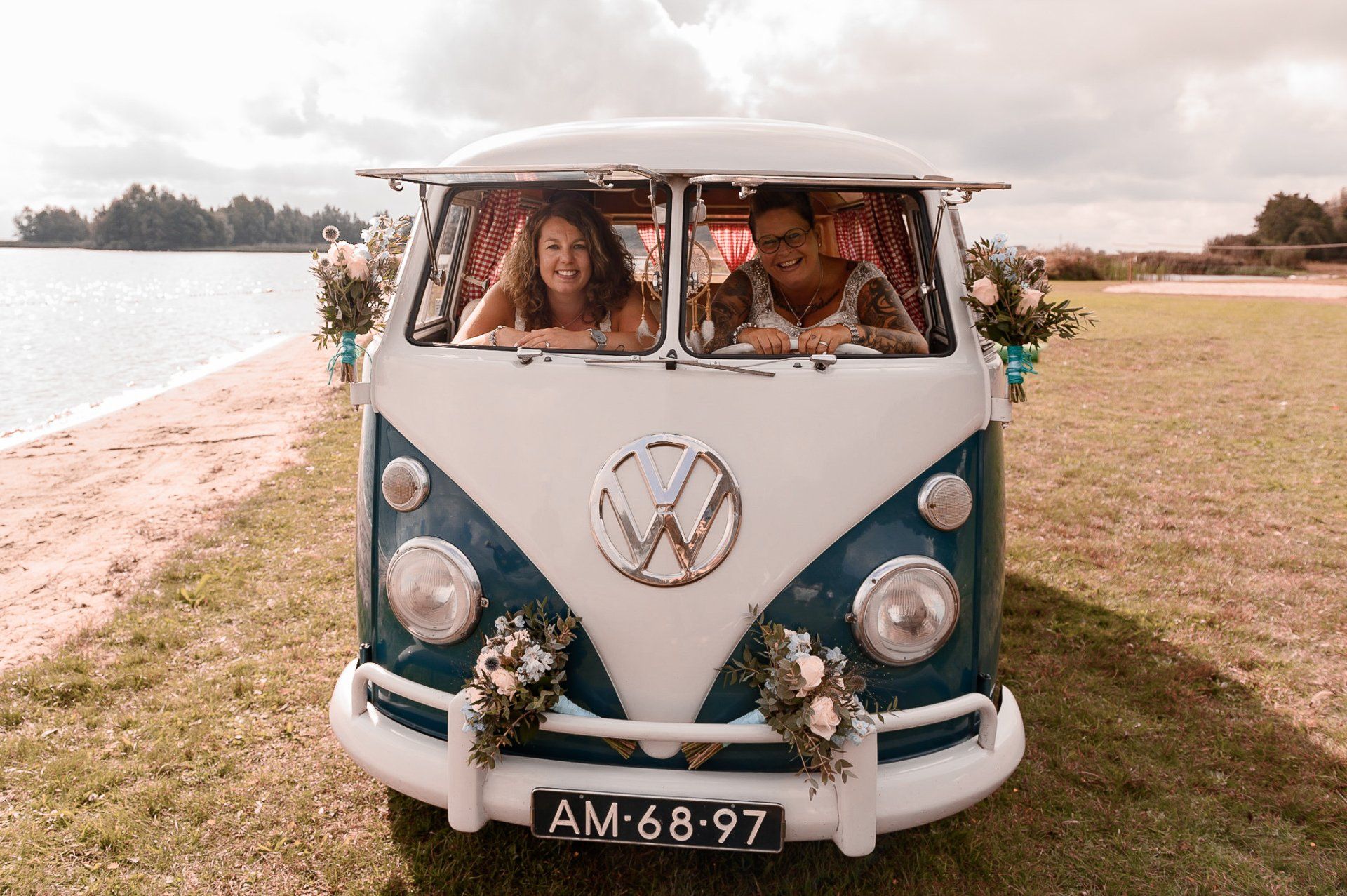 Twee mensen glimlachen vanuit een blauw-witte VW-bus met bloemversiering, vlakbij het water.
