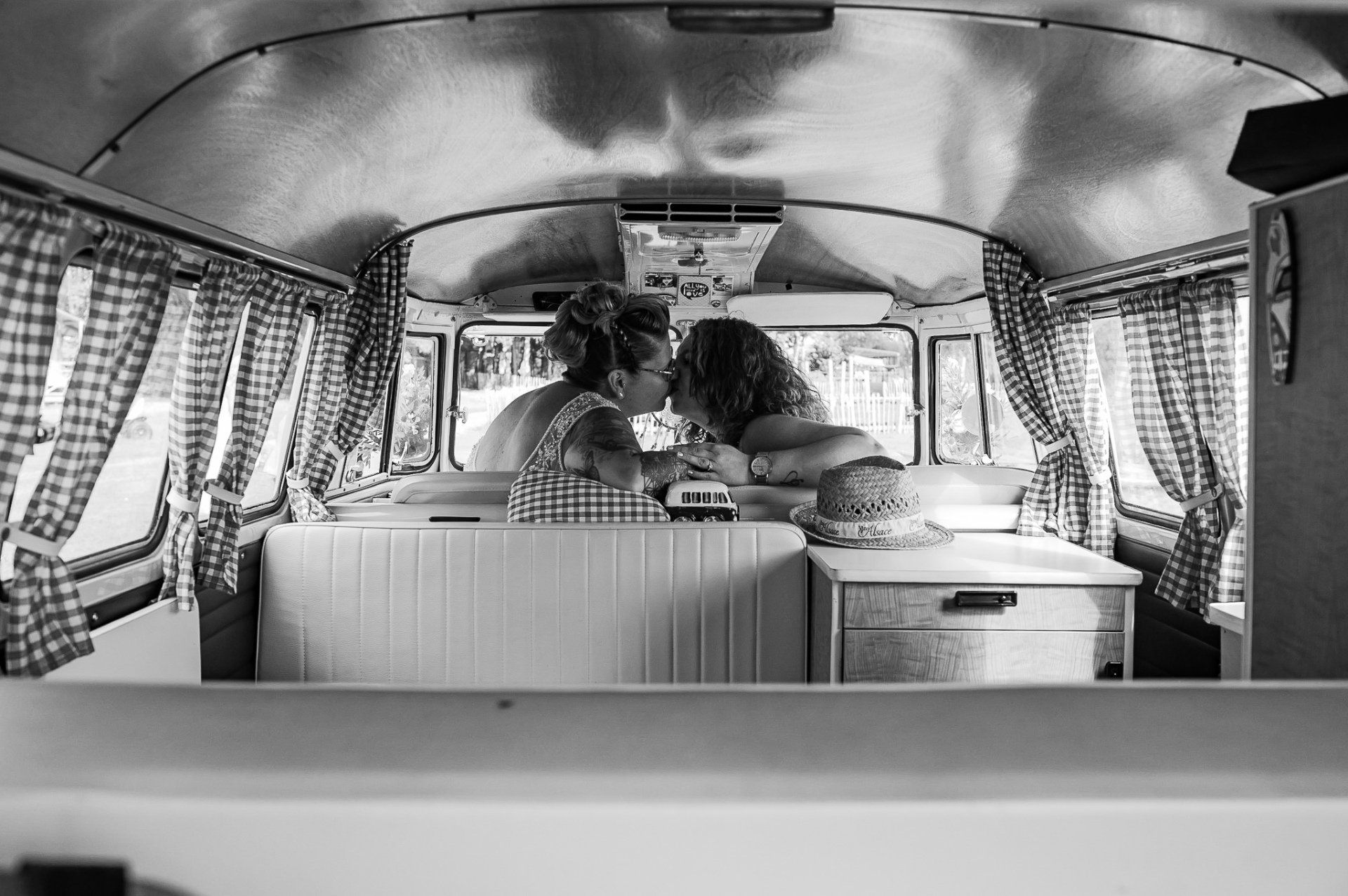 Twee mensen zoenen in een vintage camper. Geruite gordijnen omlijsten de scène.