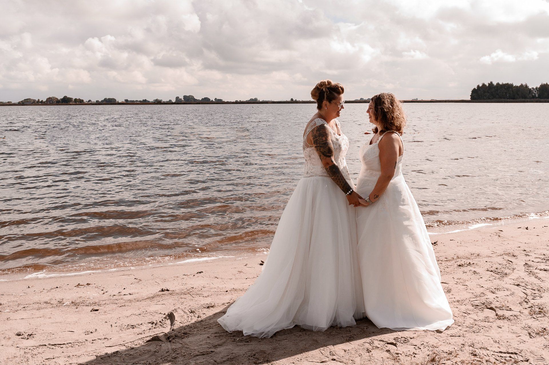 Twee vrouwen in trouwjurken staan ​​hand in hand op het strand en kijken elkaar aan.
