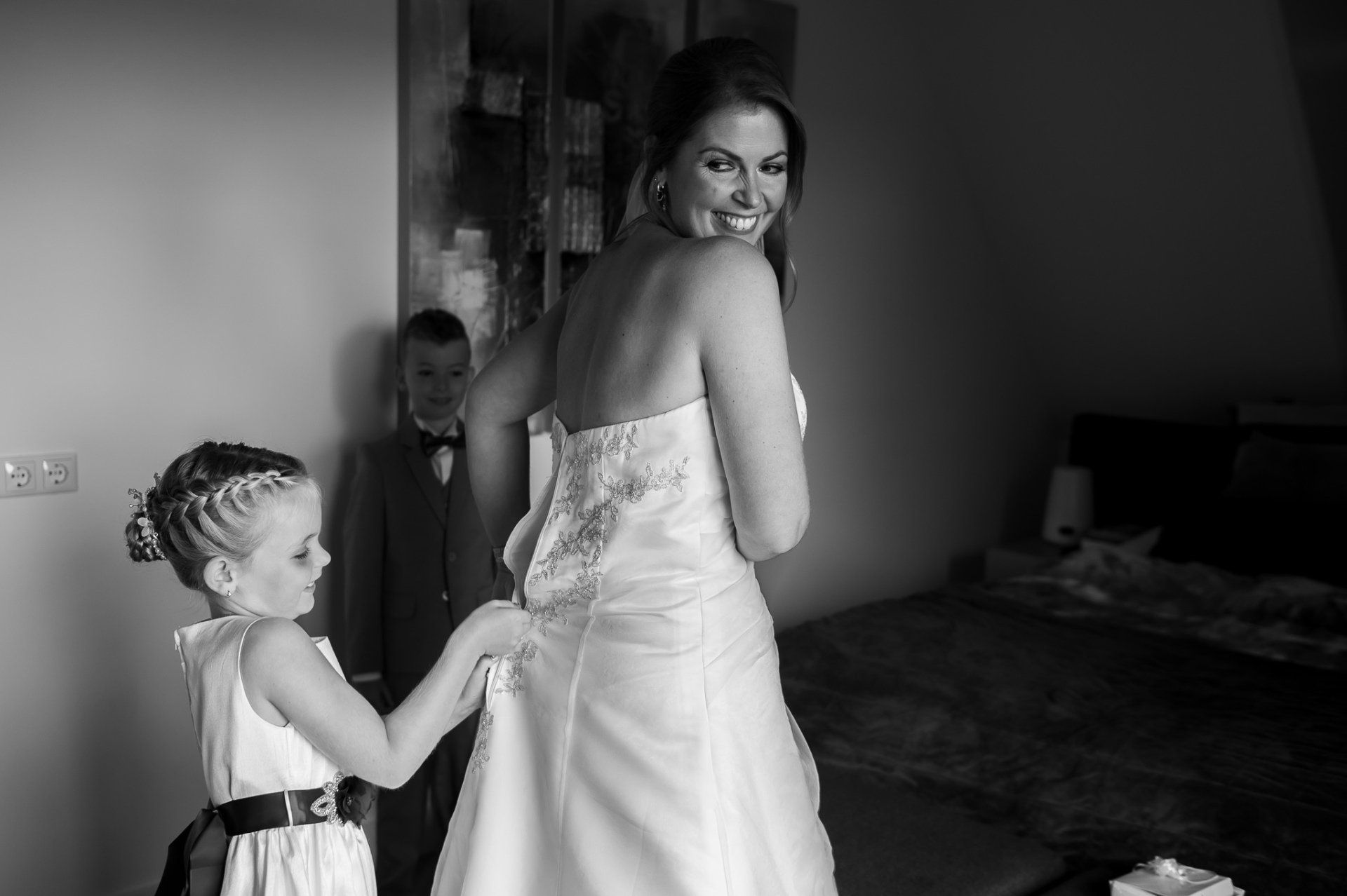Bruid in strapless jurk laat jurk aanpassen door bloemenmeisje, jongen in pak kijkt toe, raamlicht.