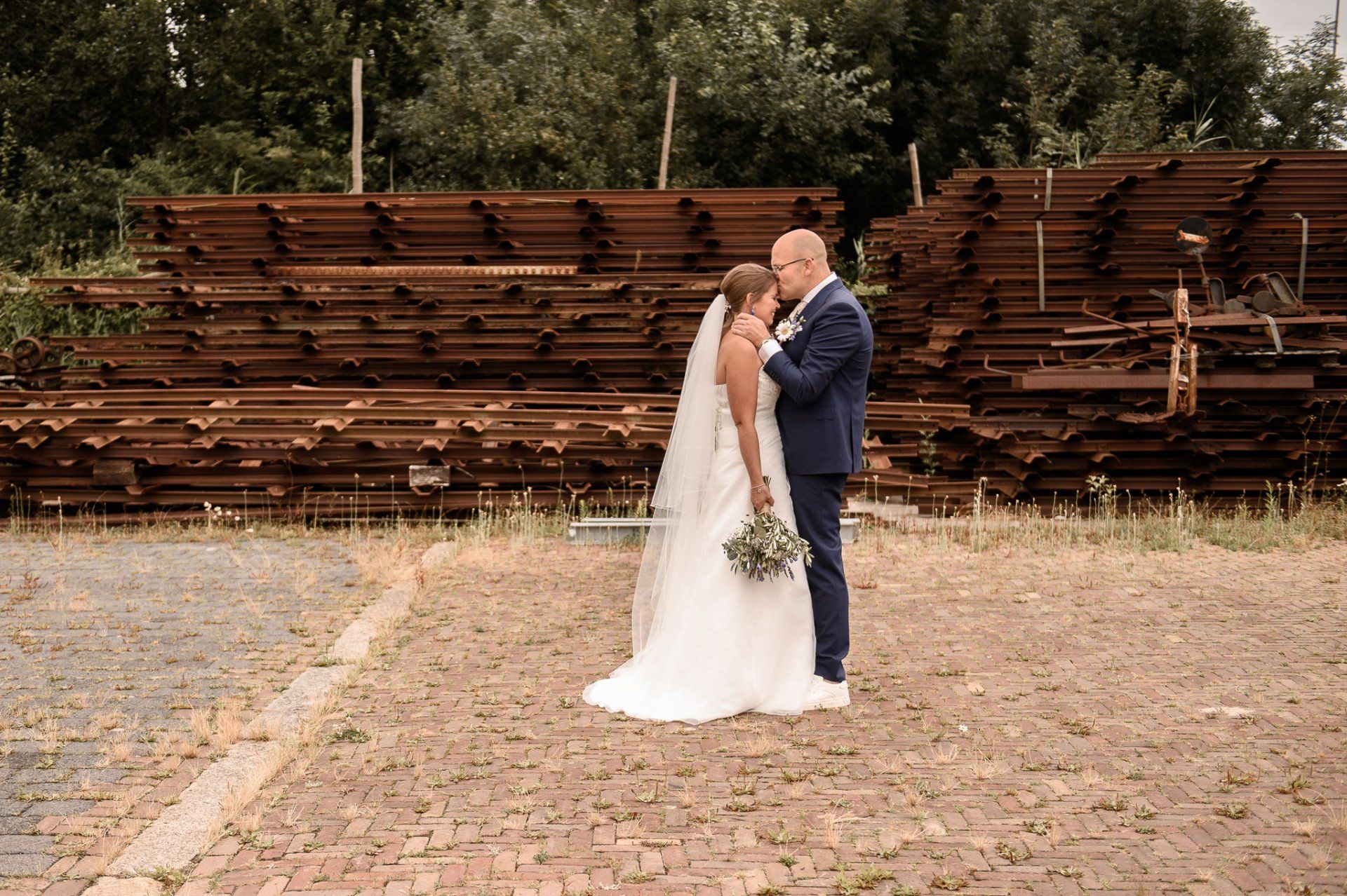 Bruid en bruidegom omhelzen elkaar buiten, vlakbij een hoop roestige spoorrails. De bruid draagt ​​een witte jurk en sluier. De bruidegom draagt ​​een blauw pak.