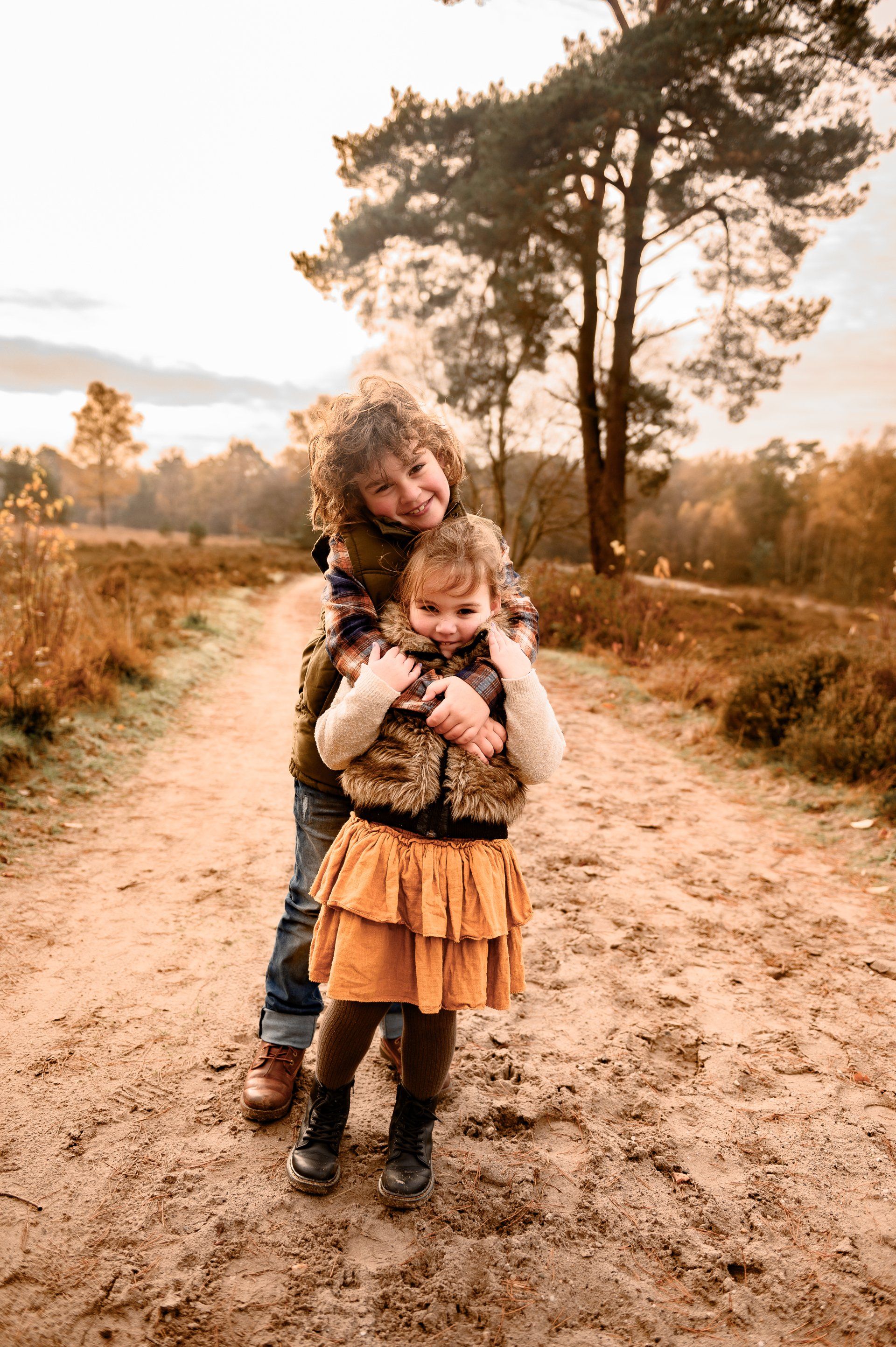 Twee kinderen omhelzen elkaar op een zandpad in een veld. Het langere kind omhelst het kortere kind. Herfstkleuren.