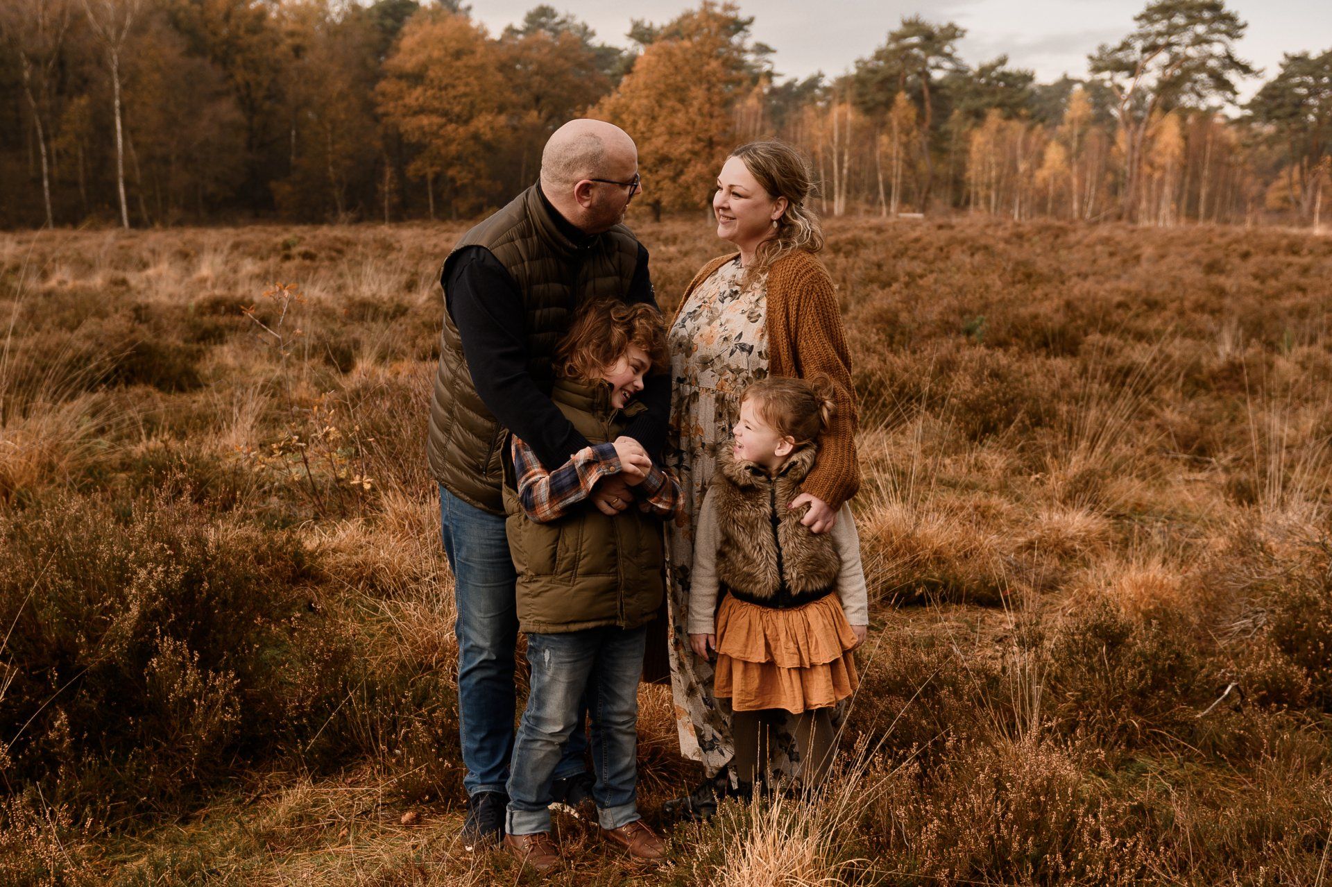 Gezin in een buitenveld, moeder zwanger, kinderen omarmend. Bruine en groene tinten, herfstsetting.