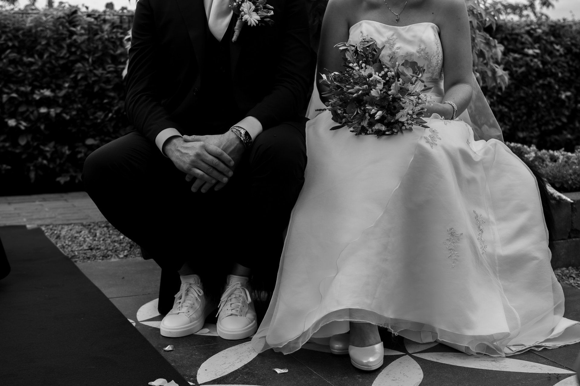 De bruid in de witte jurk zit naast de man in het pak en de sportschoenen. Ze zijn buiten.
