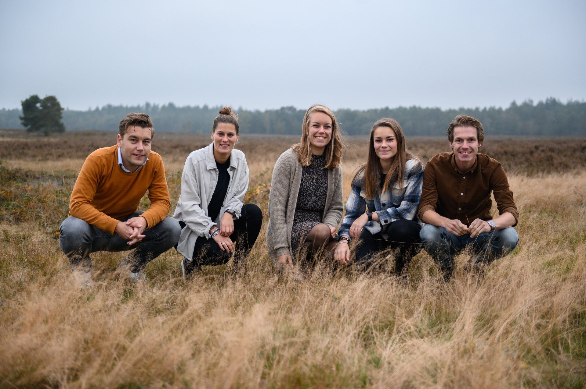 Vijf mensen zaten gehurkt in een veld en glimlachten. Het was een bewolkte dag met bruin gras en bomen op de achtergrond.
