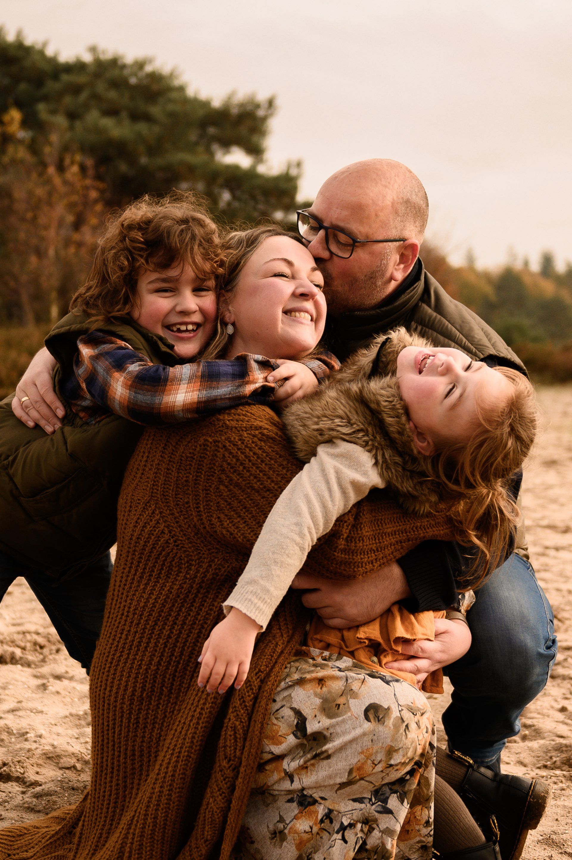 Een gezin van vier personen omhelst elkaar buiten, twee kinderen glimlachen, volwassenen omhelzen elkaar.