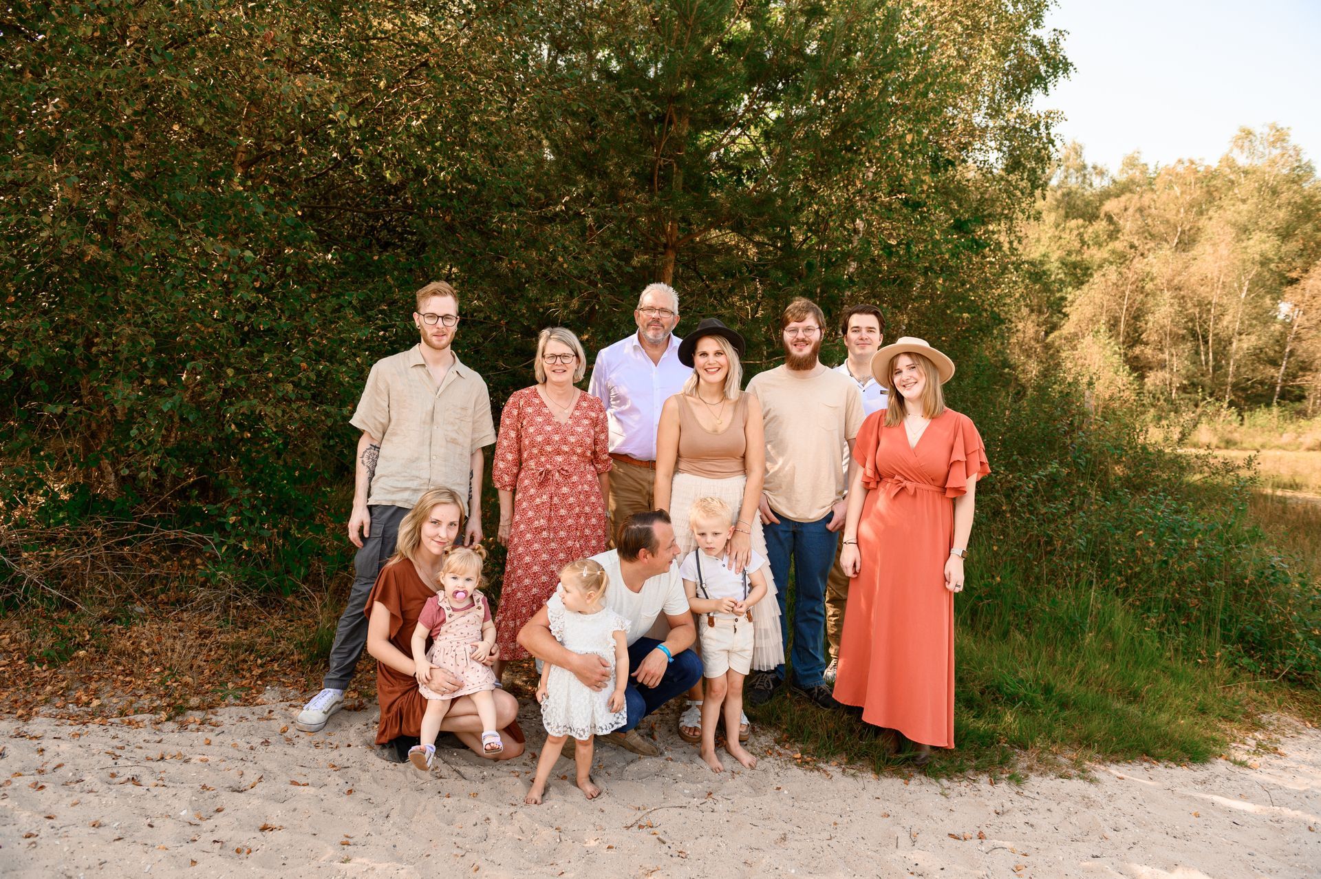 Familieportret in de buitenlucht, poserend op een zandpad. De mensen dragen neutrale kleuren tegen een achtergrond van groen gebladerte.