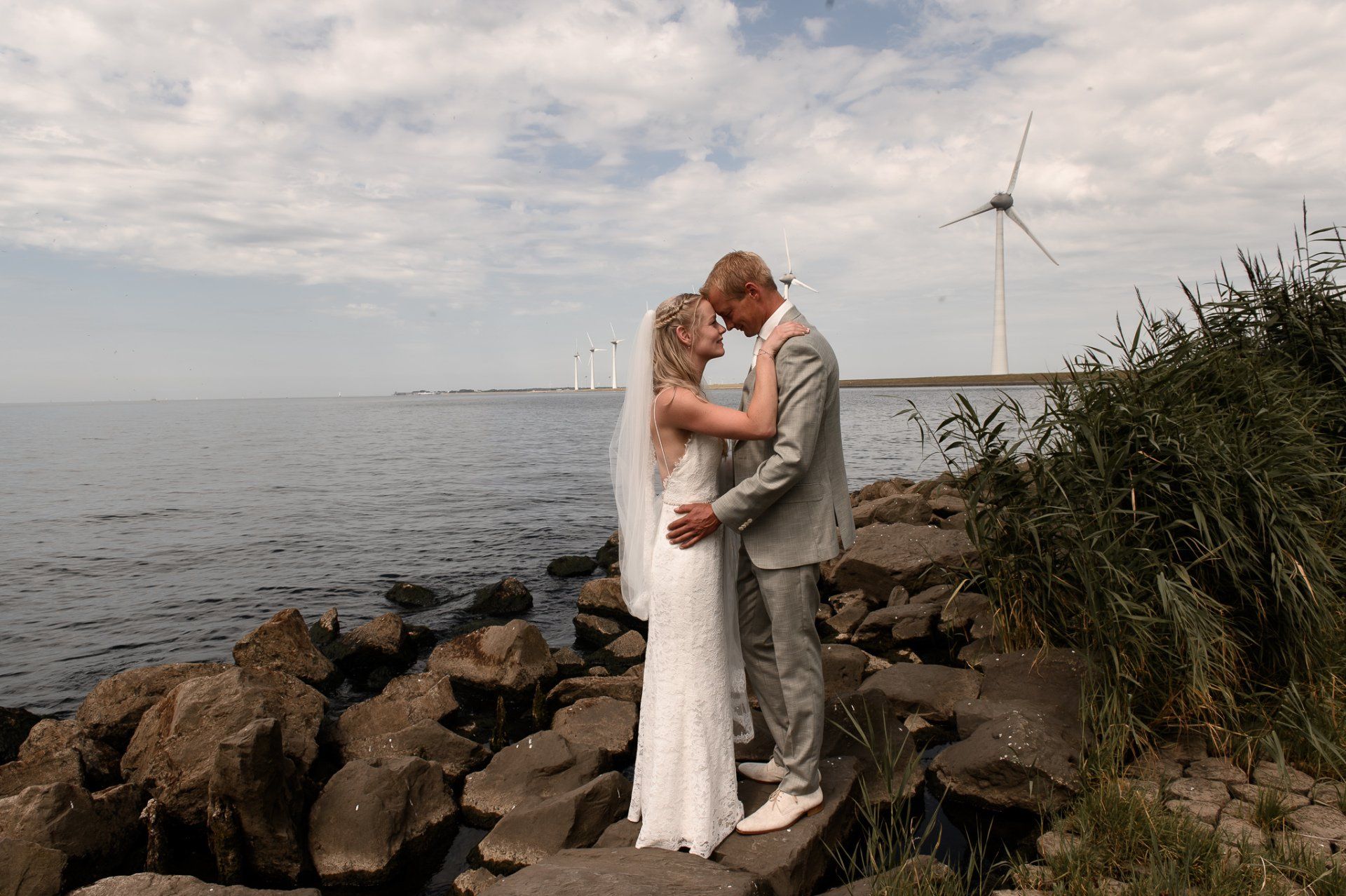 Bruid en bruidegom omhelzen elkaar aan het water op hun trouwdag, met windturbines in de verte.
