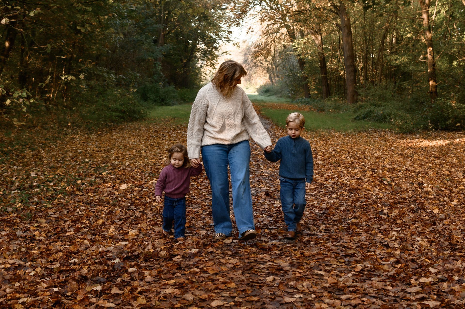 Een vrouw loopt hand in hand met twee jonge kinderen over een met bladeren bedekt pad in een bosrijk gebied.