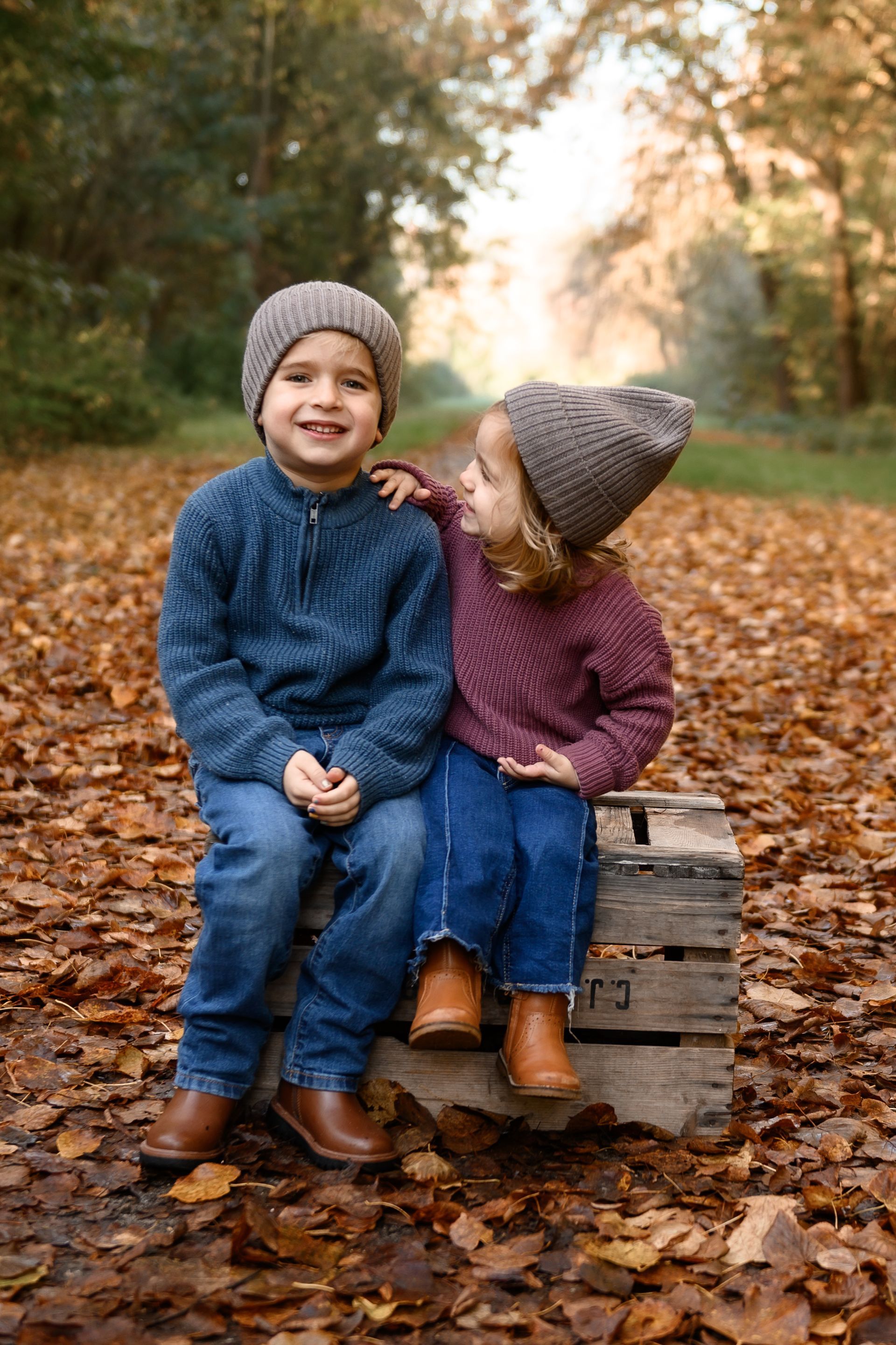 Twee kinderen zitten op een houten kist op een met bladeren bedekt pad; de jongen lacht, het meisje raakt zijn schouder aan, ze dragen alle drie gebreide mutsen.
