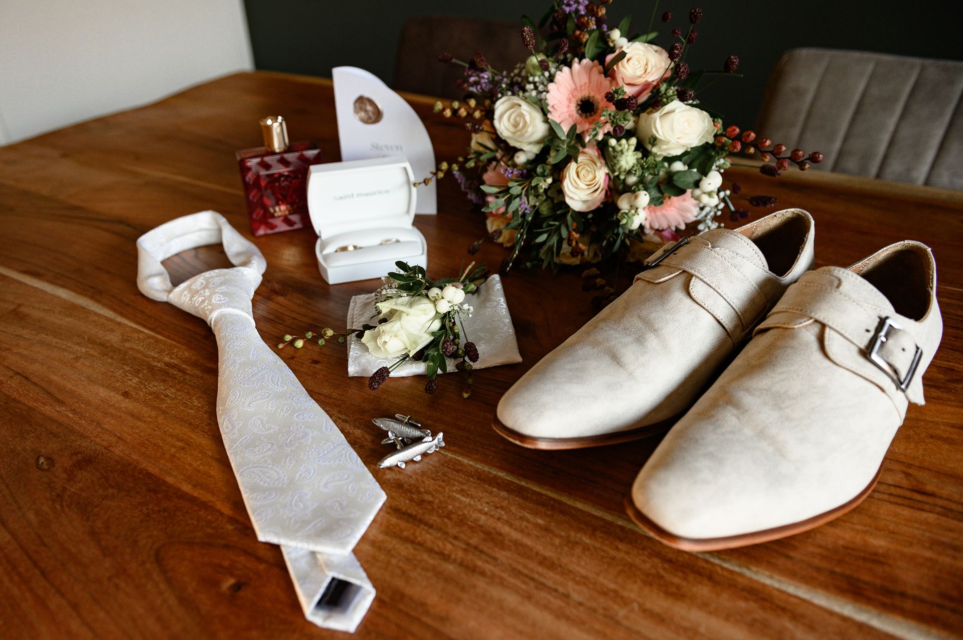 De opstelling voor de trouwdag: stropdas, schoenen, ringen, bloemen, parfum en manchetknopen op een houten tafel.