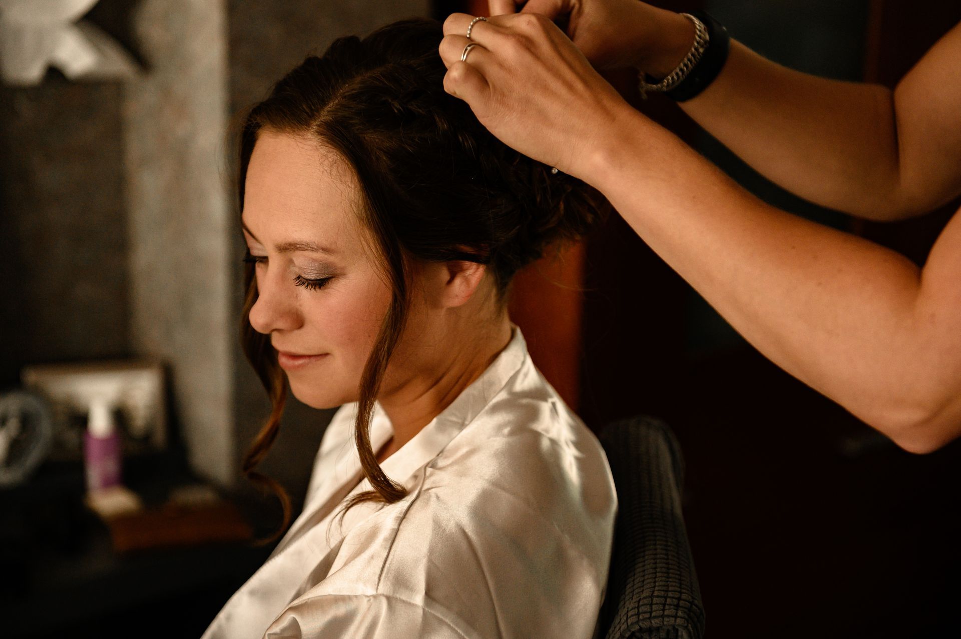 Een vrouw krijgt haar haar gestyled; de handen van de stylist zijn zichtbaar. Binnen draagt ​​ze een witte ochtendjas, heeft ze haar ogen gesloten en een lichte glimlach.