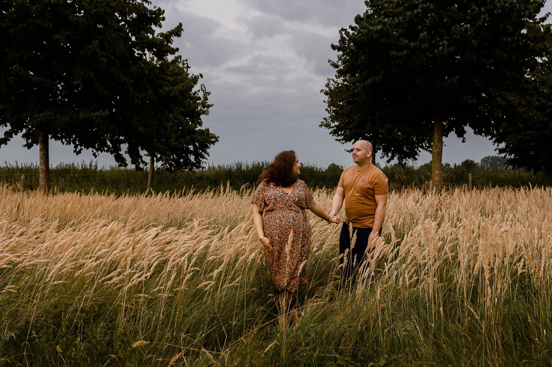 Een stel loopt hand in hand door het hoge gras in een veld tussen twee bomen. Bewolkte lucht.