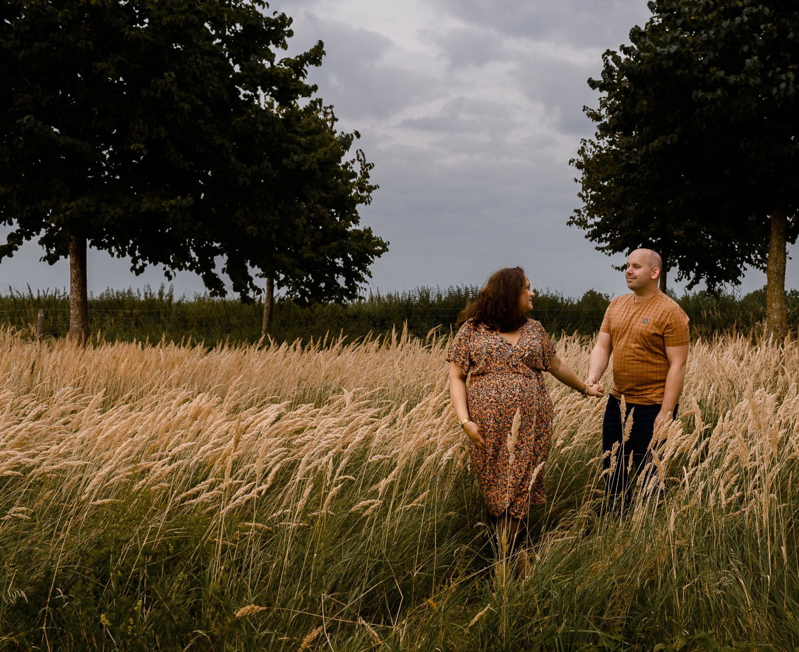 Een zwanger stel loopt hand in hand door een veld met hoog gras, donkere bomen en een bewolkte hemel.