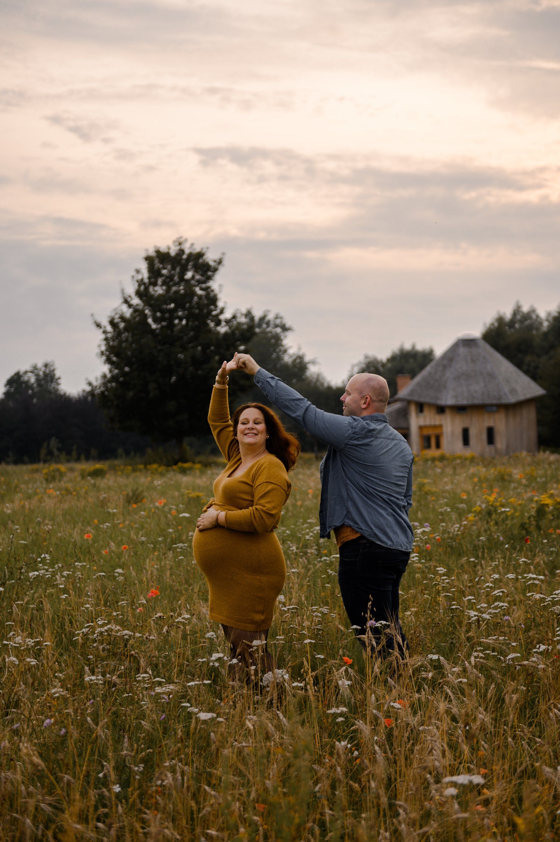 Een zwangere vrouw danst met een man in een veld vol wilde bloemen bij zonsondergang.