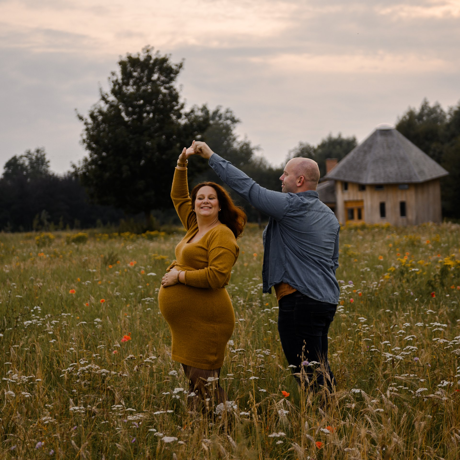 Een zwangere vrouw en een man dansen in een veld vol wilde bloemen bij een houten gebouw. ​​Warme tinten.