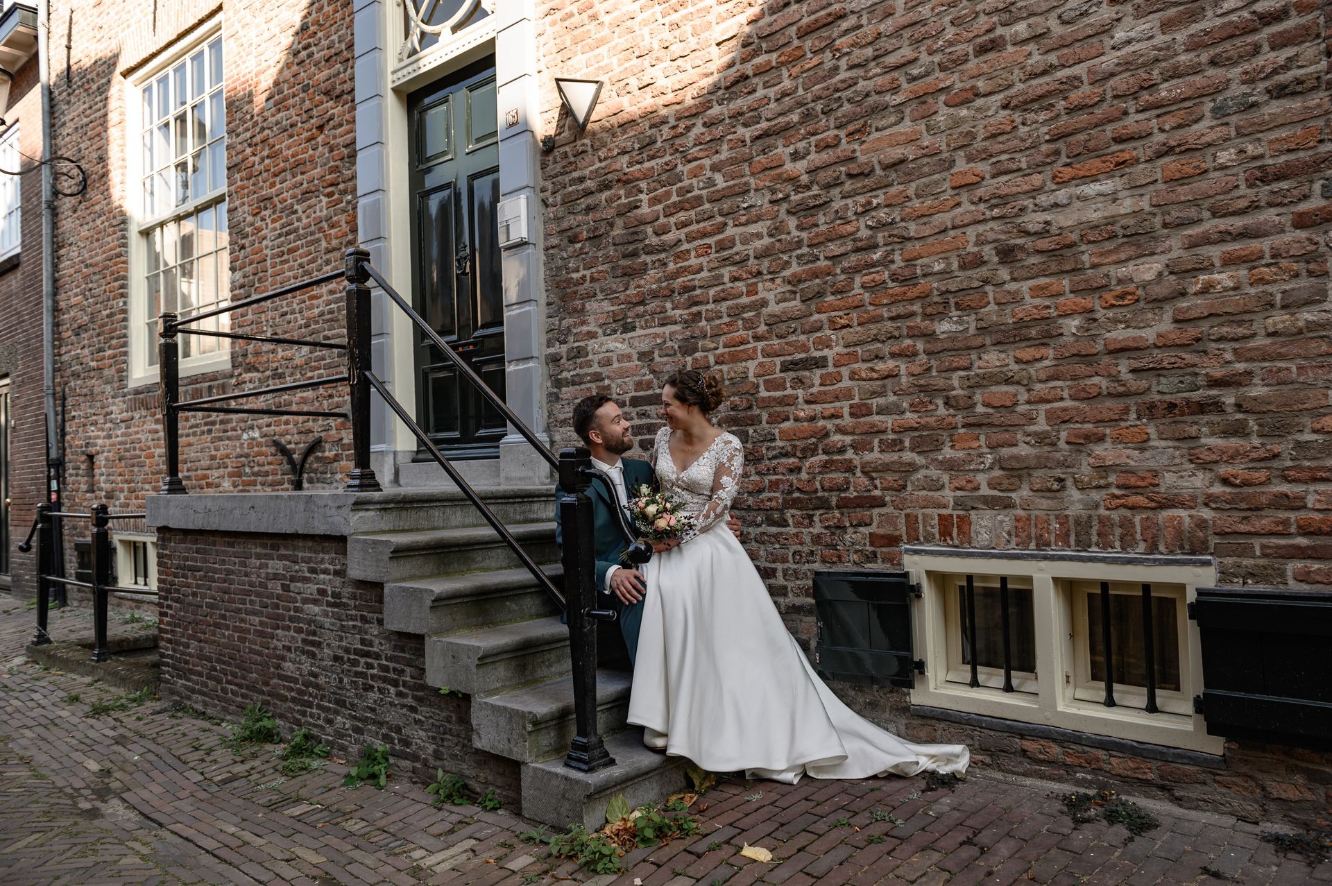Een bruidspaar staat lachend op de trappen voor een bakstenen gebouw met een zwarte deur.