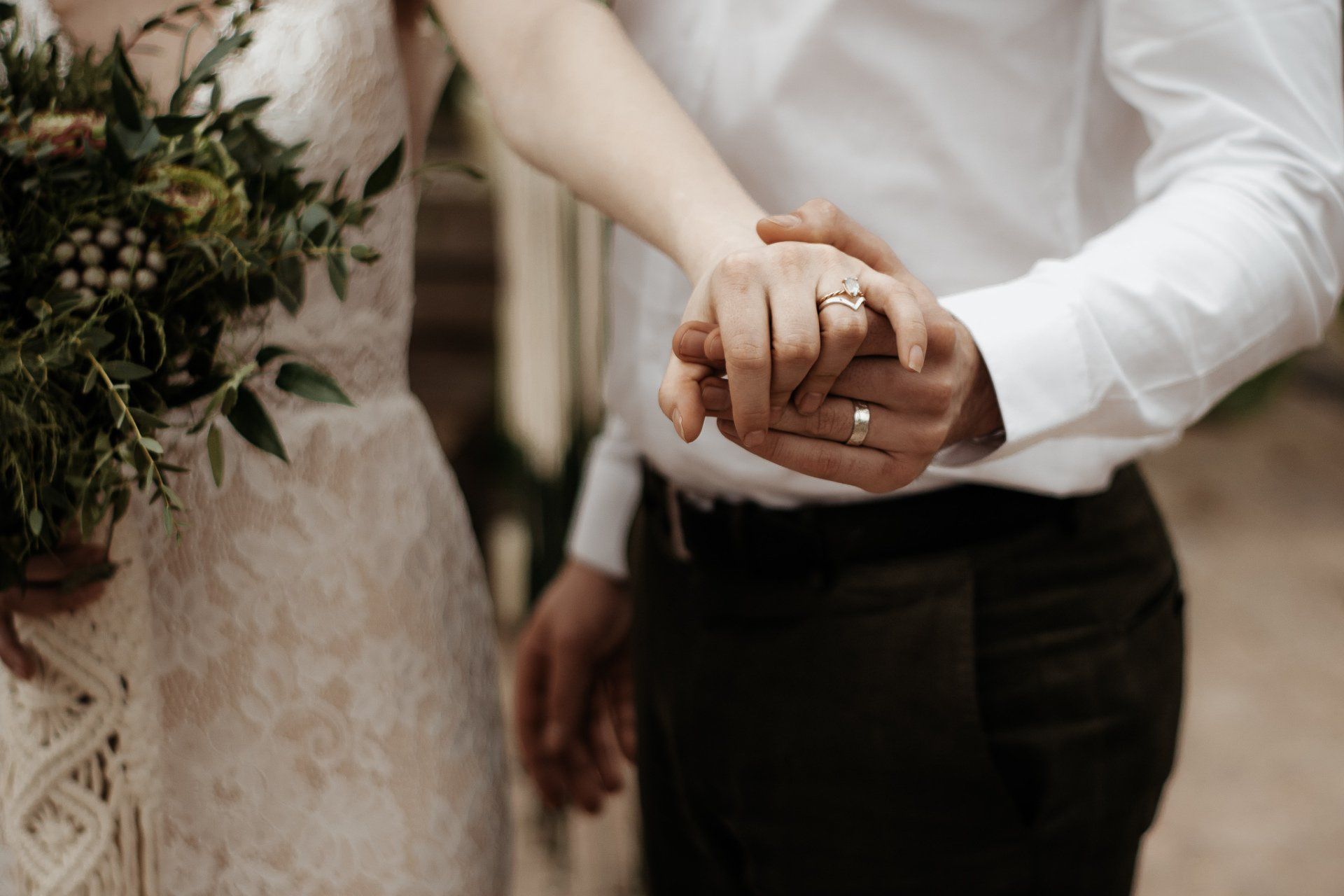 Bruidspaar hand in hand, ringen zichtbaar. Vrouw in kanten jurk houdt boeket vast.