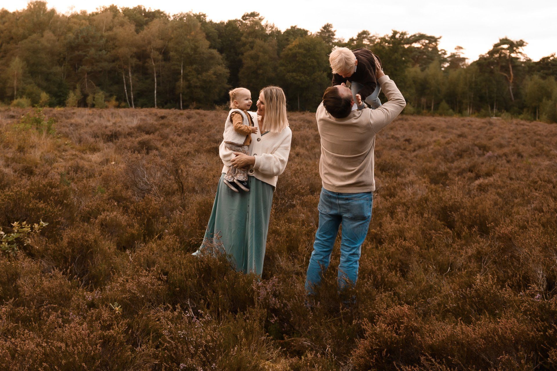 Een gezin speelt in een veld met paarse bloemen. Ouders houden hun kinderen vast en gooien ze in de lucht, terwijl ze glimlachen. Op de achtergrond zijn bomen te zien.