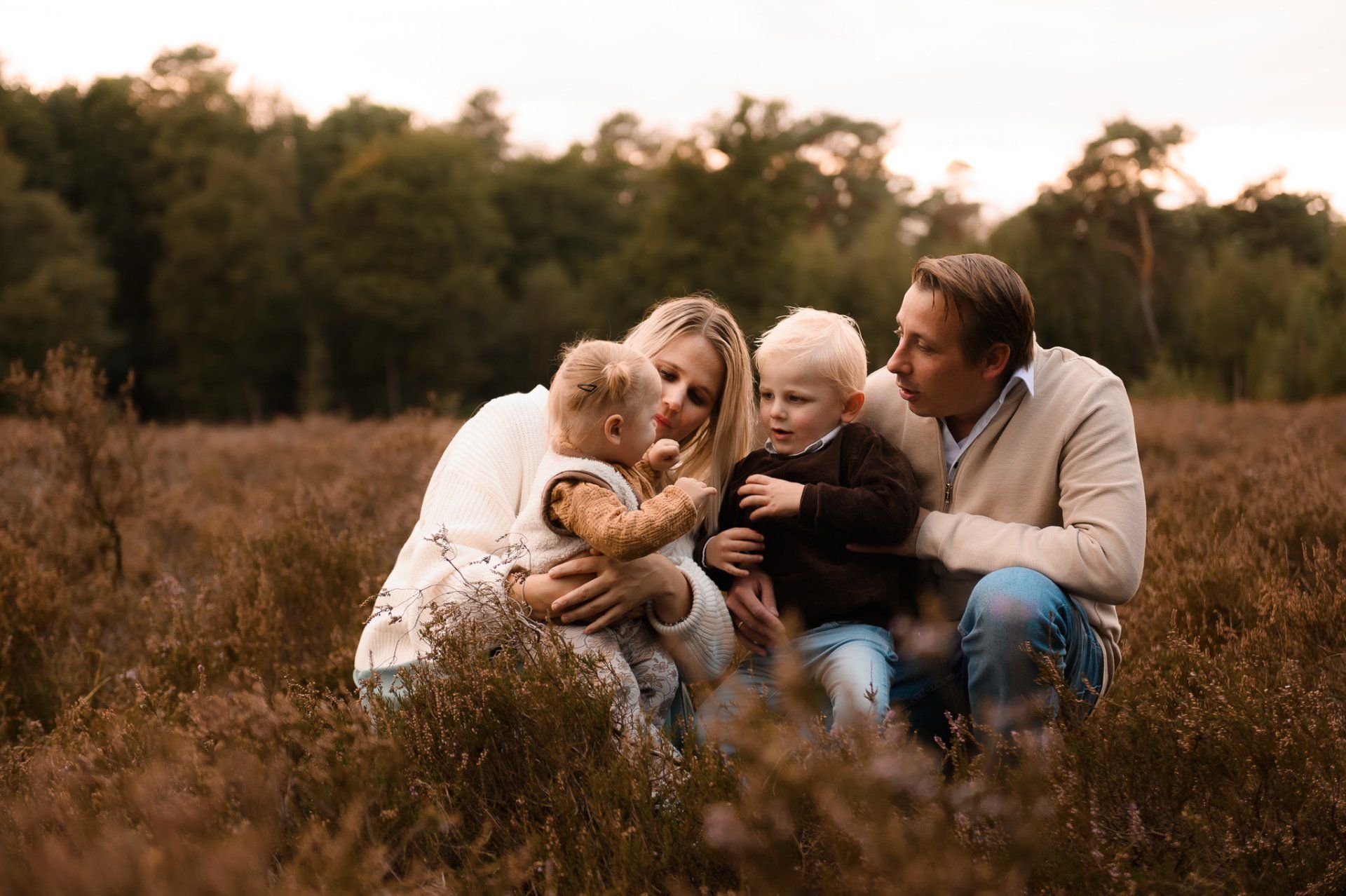 Een gezin van vier in een veld, moeder met kind op de arm, ander kind en vader in de buurt, allen lachend.
