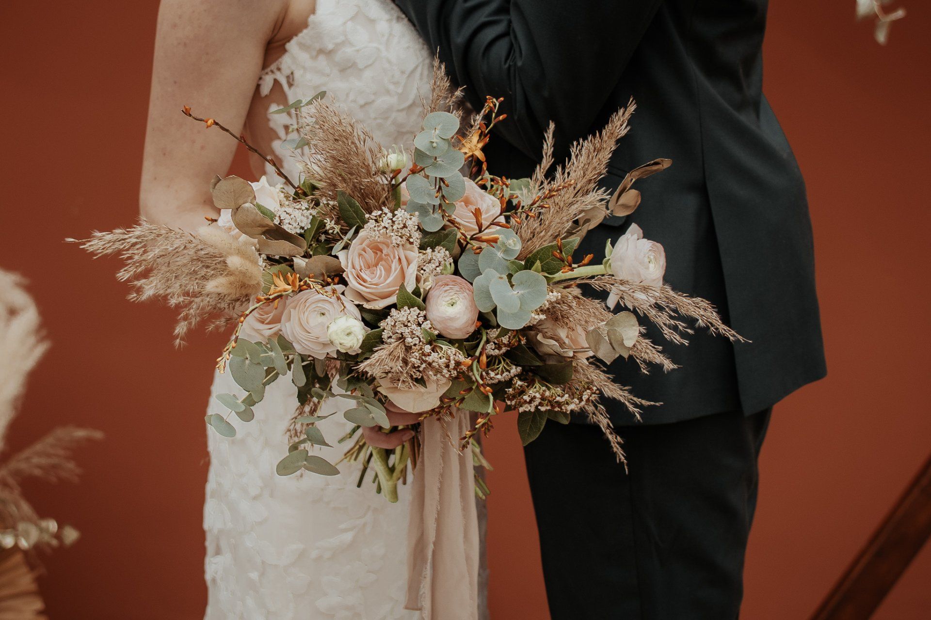 Bruid en bruidegom omhelzen elkaar en houden een boeket roze en beige bloemen vast, tegen een roestbruine achtergrond.