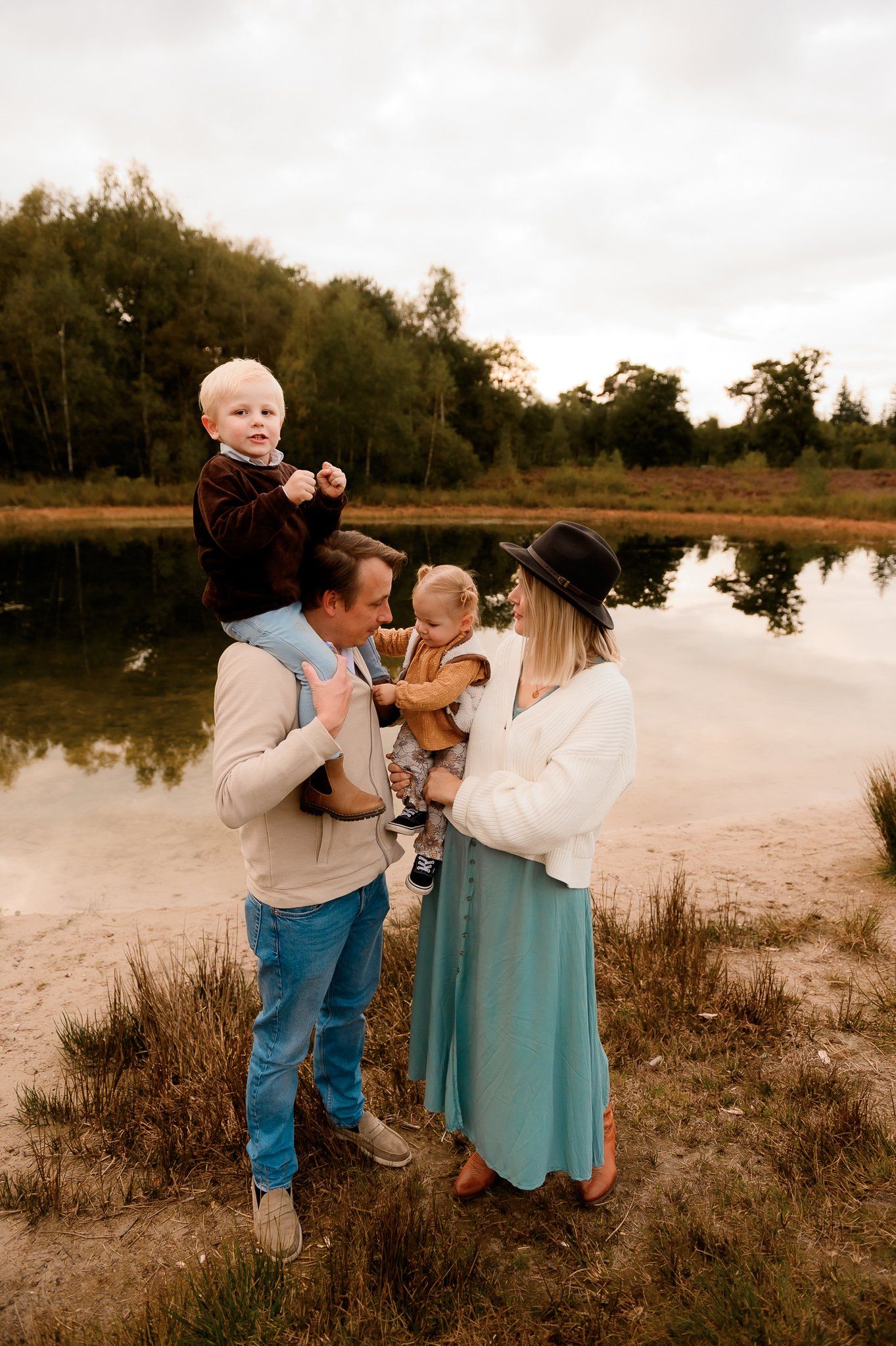 Een gezin aan een meer: ​​een man draagt ​​een kind op zijn schouders, een vrouw houdt een peuter vast, allen lachend en in vrijetijdskleding.