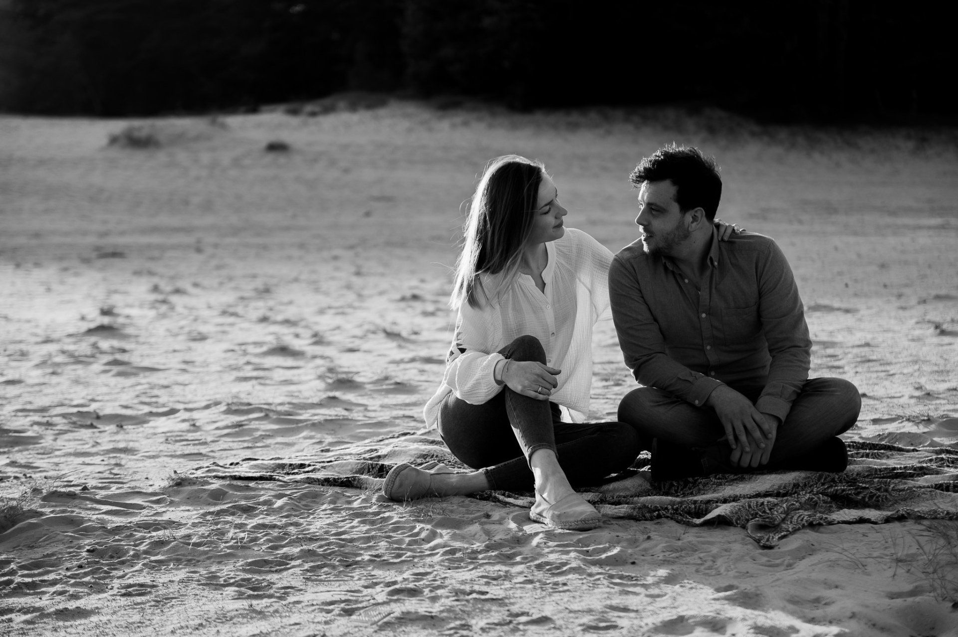 Een stel zit op het strand en kijkt elkaar aan. Ze zitten op een deken, de vrouw heeft haar been omhoog. Zwart-witfoto.