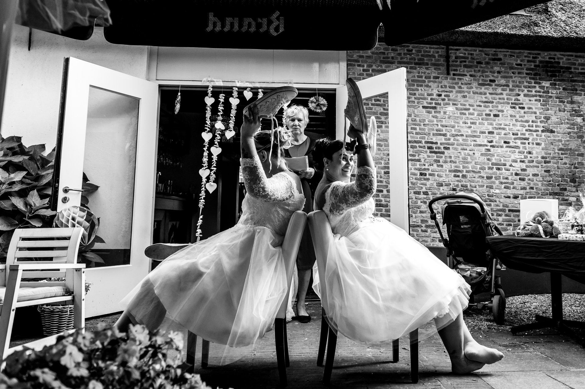 Twee vrouwen in trouwjurken, arm in arm, tillen stoelen op en staan ​​tegenover elkaar met een ambtenaar van de burgerlijke stand voor een open deuropening.