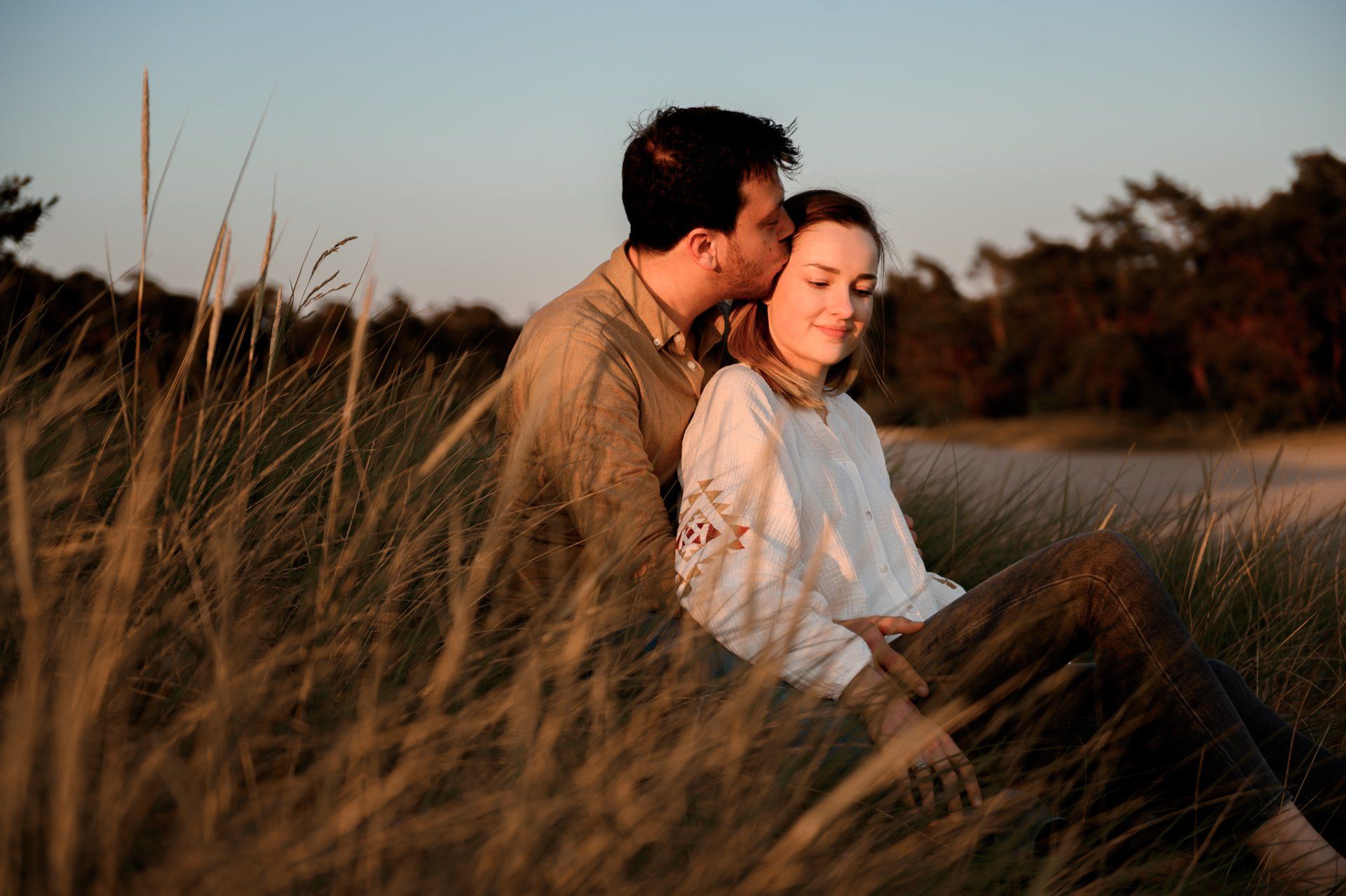 Een man kust het voorhoofd van een vrouw terwijl ze samen in het hoge gras zitten, verlicht door de warme zon.
