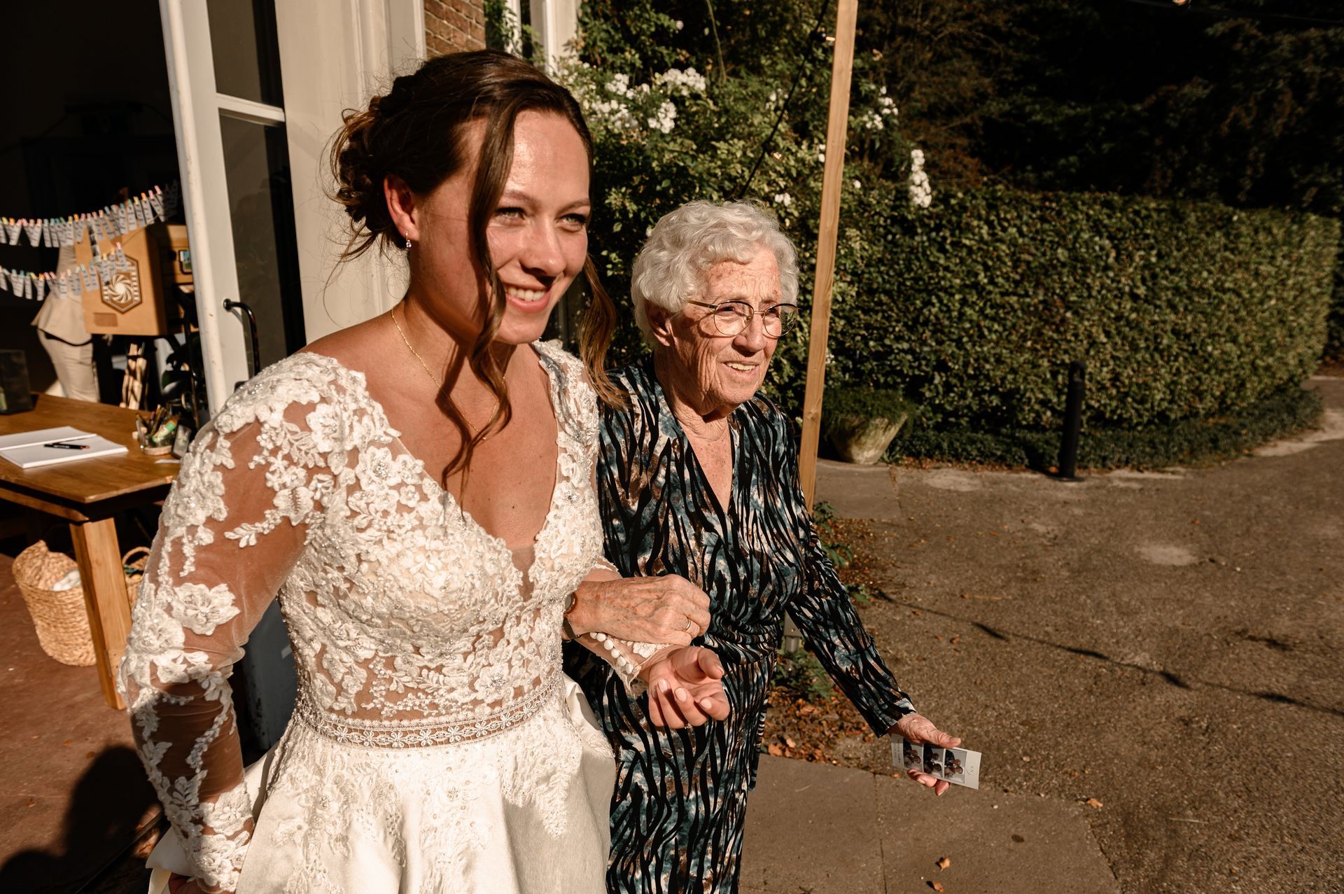 Bruid in kanten trouwjurk lacht terwijl ze arm in arm met oudere vrouw buiten loopt.