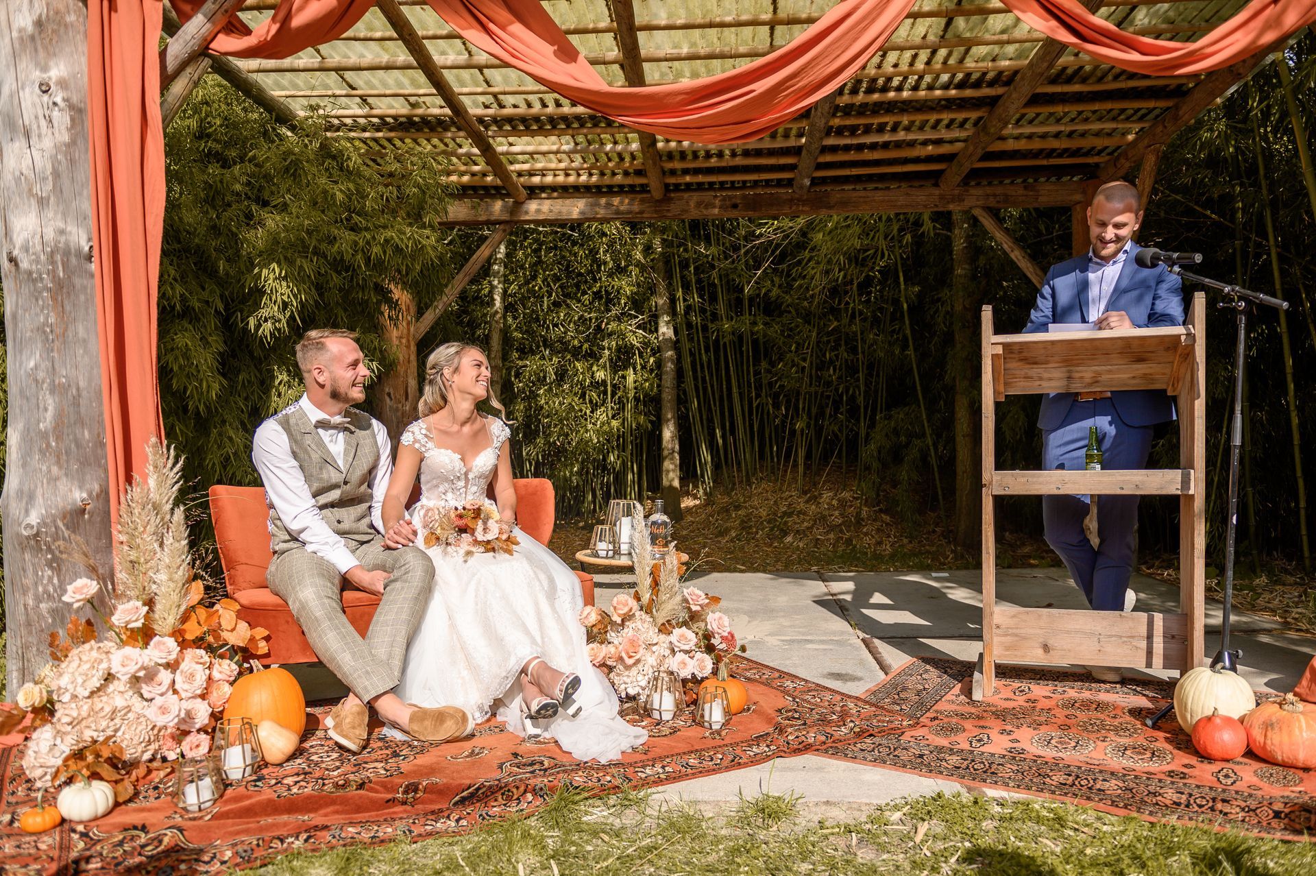 Huwelijksceremonie: Bruid en bruidegroom zitten glimlachend te luisteren naar de ambtenaar van de burgerlijke stand onder een met oranje draperieën en herfstdecoratie versierde pergola.