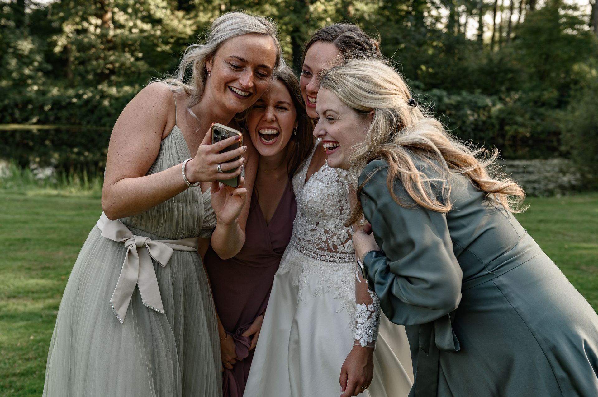 Vijf vrouwen, waaronder een bruid in een witte jurk, lachen terwijl ze buiten naar een telefoon kijken.