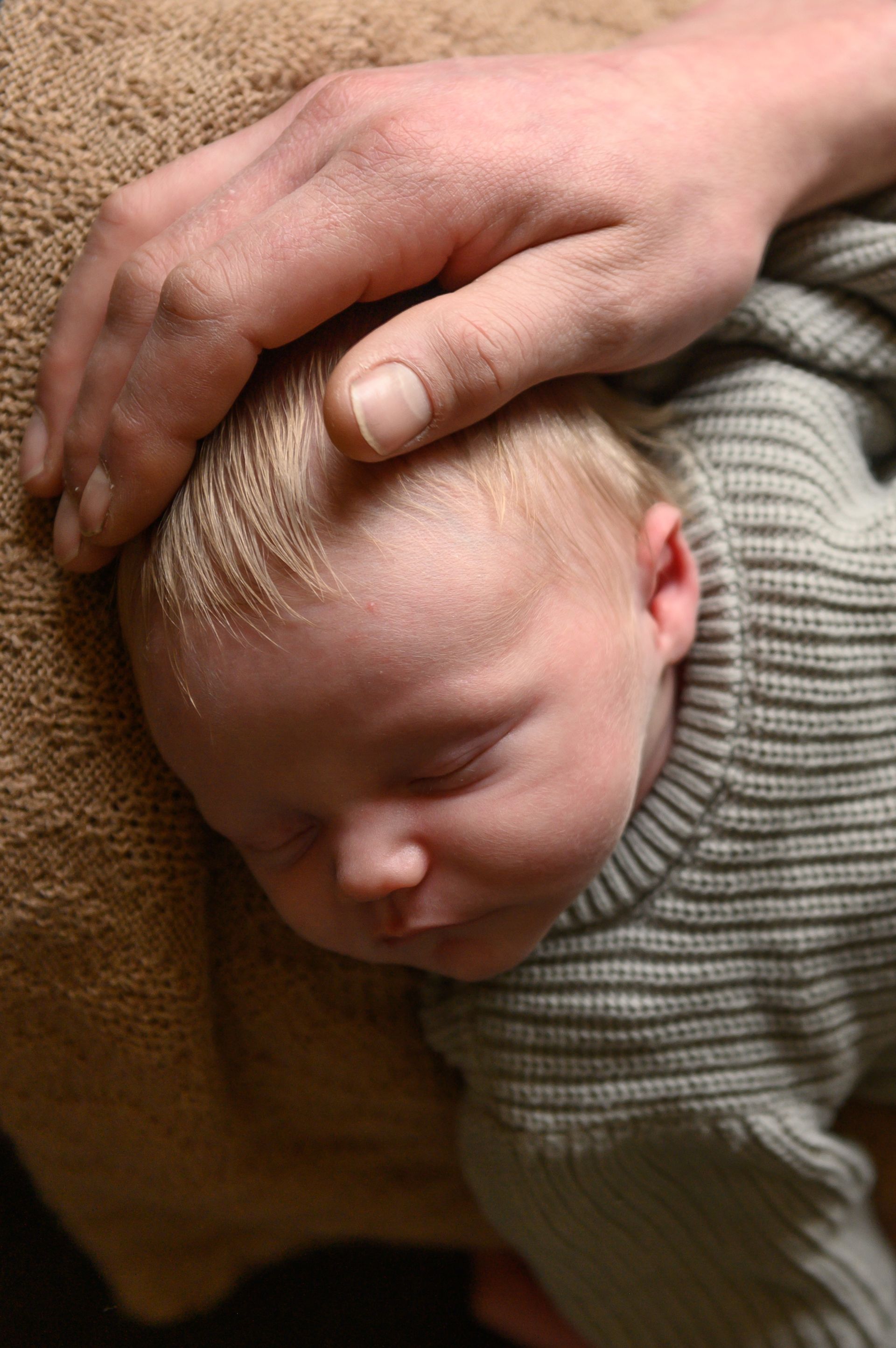 Een hand rust zachtjes op het hoofdje van een slapende baby die een grijs gestreept truitje draagt.