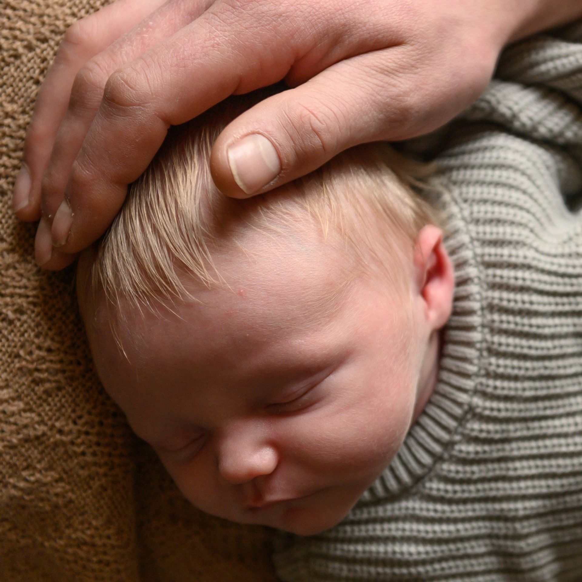 Een hand rust zachtjes op het hoofdje van een slapende baby. De baby draagt een grijs truitje en ligt op een bruine ondergrond.