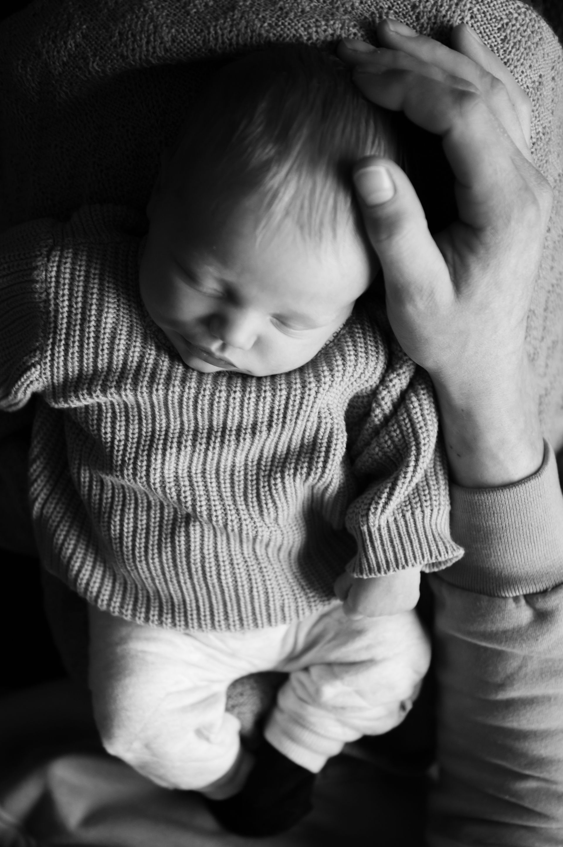 Pasgeboren baby, gekleed in een gebreid truitje en een witte broek, wordt in iemands armen gehouden. Een hand rust zachtjes op het hoofdje van de baby.