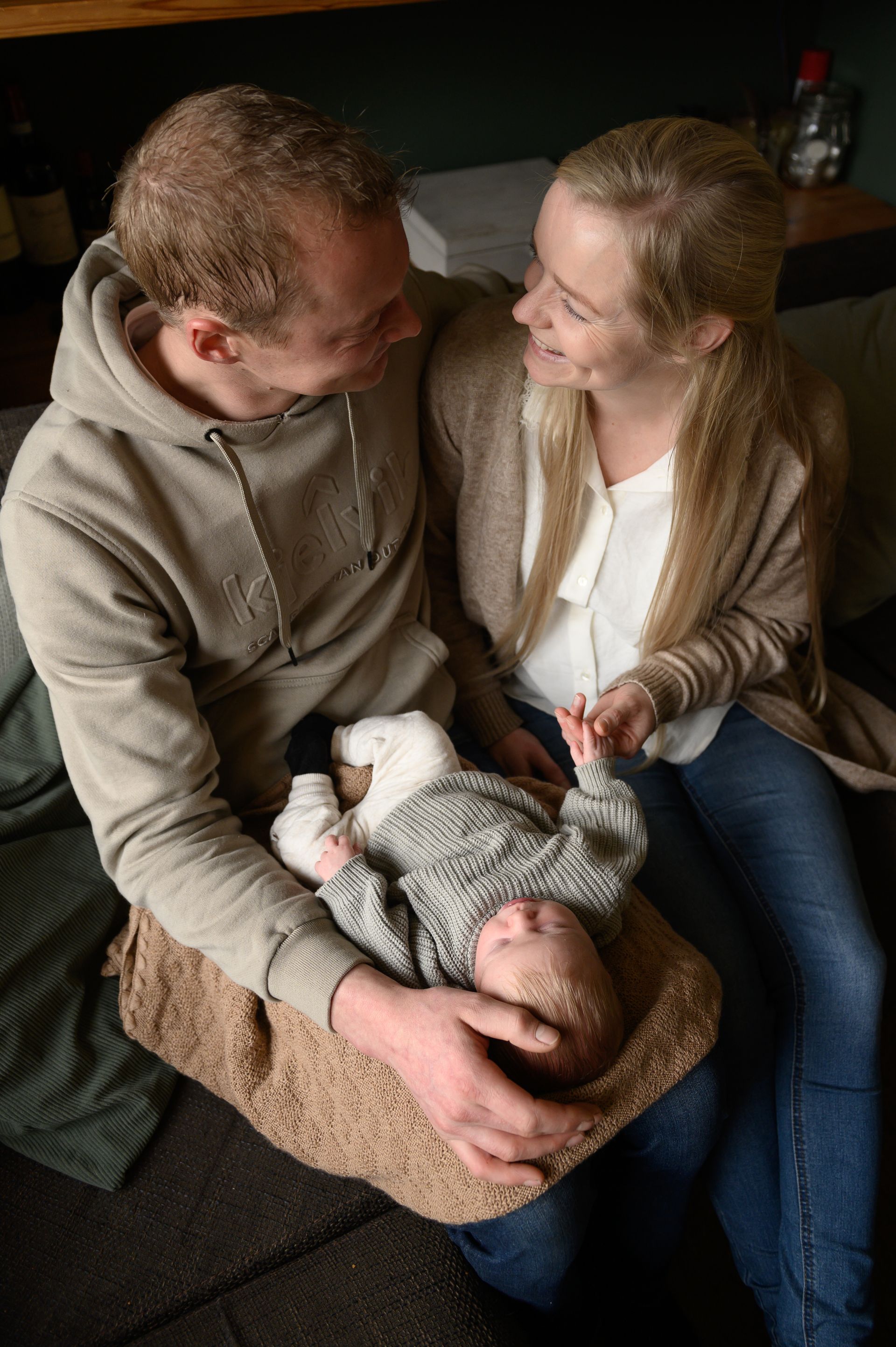 Een stel lacht naar elkaar terwijl ze een baby vasthouden op een bank. De baby draagt ​​een grijs pakje.