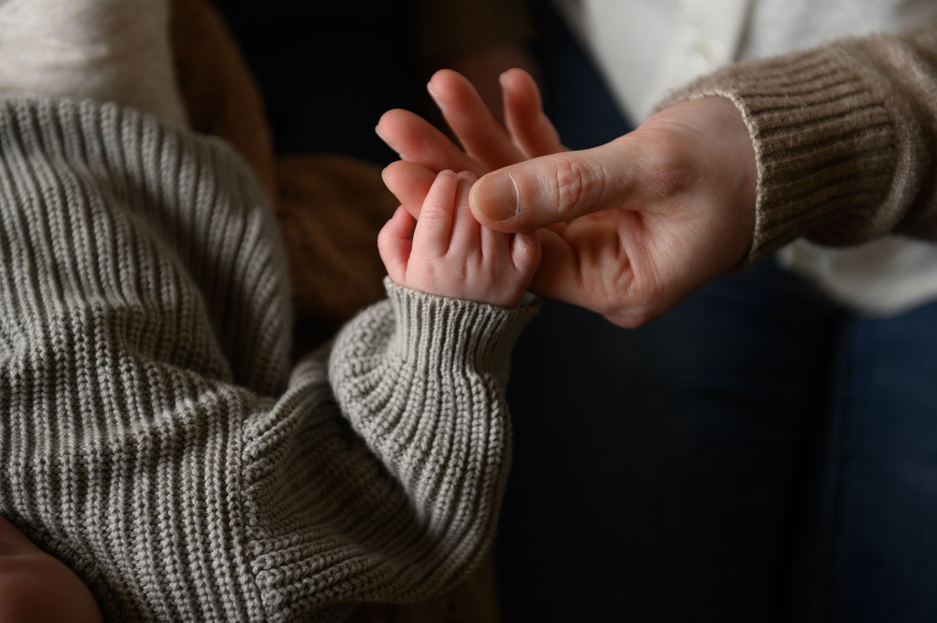 Een volwassen hand houdt de hand van een baby vast; beiden dragen gebreide truien.