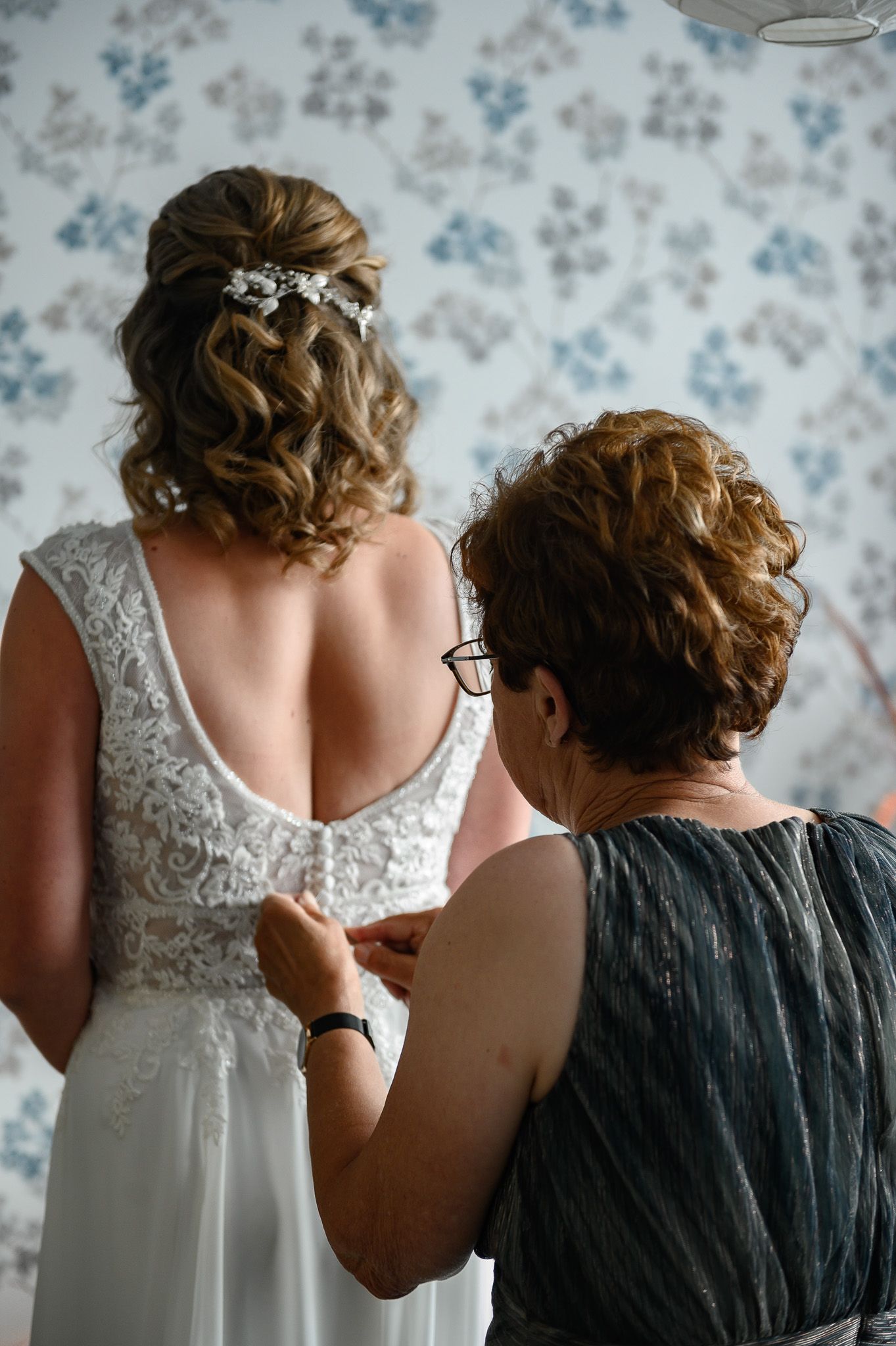 Een vrouw in een trouwjurk, met haar rug naar de camera, wordt binnenshuis door een andere vrouw dichtgeknoopt.