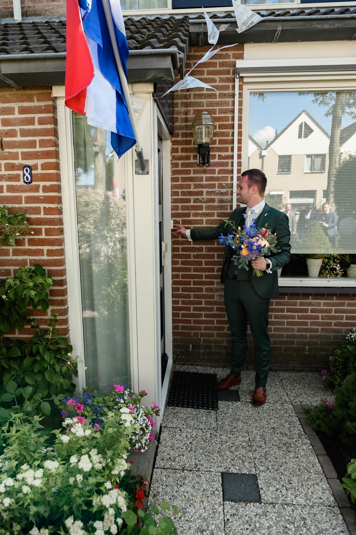 Een man in een groen pak belt aan bij een bakstenen huis. Hij houdt bloemen vast en er hangt een Nederlandse vlag boven de deur.