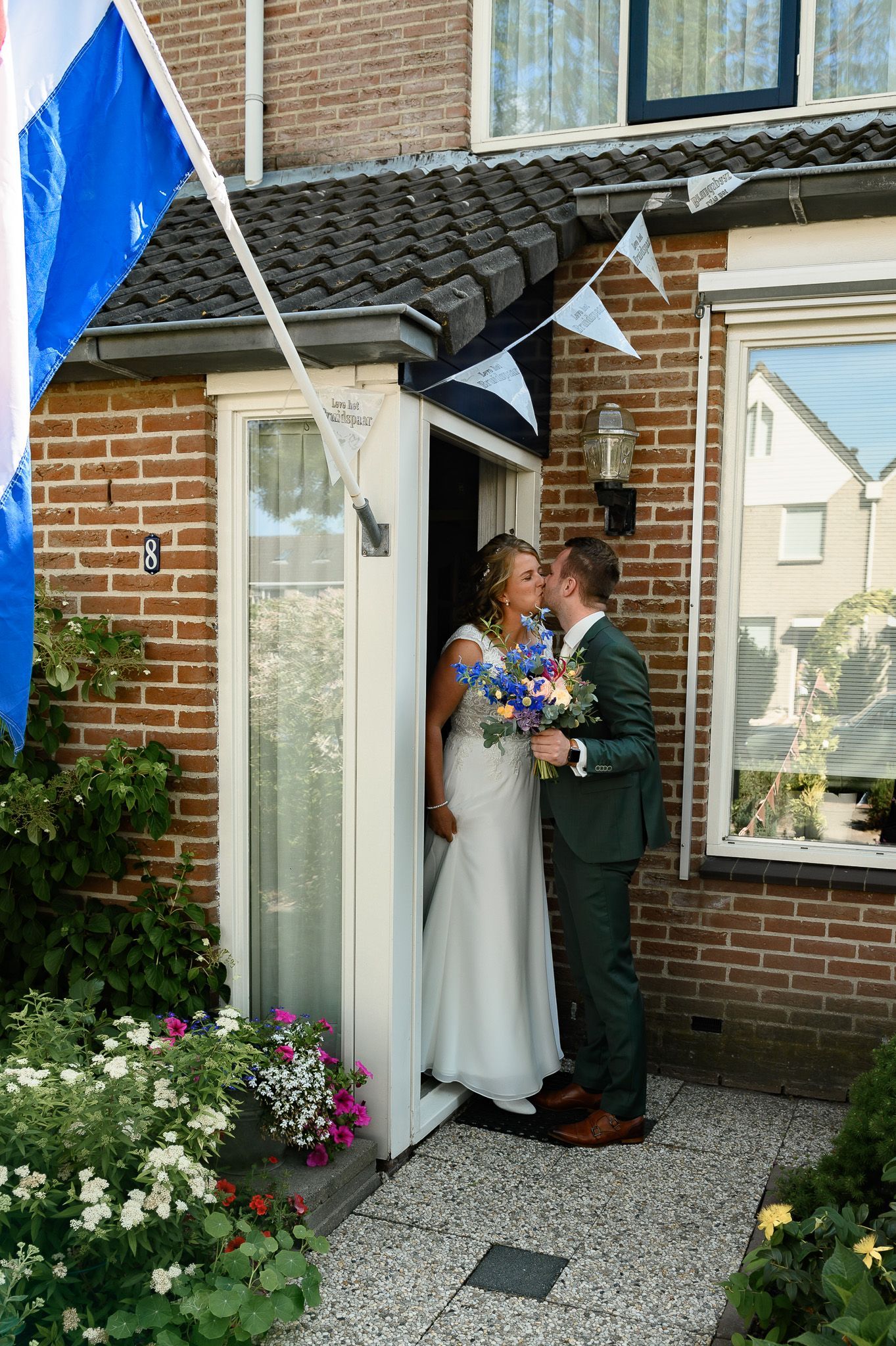 Bruid en bruidegom kussen elkaar in de deuropening van een feestelijk versierd bakstenen huis.