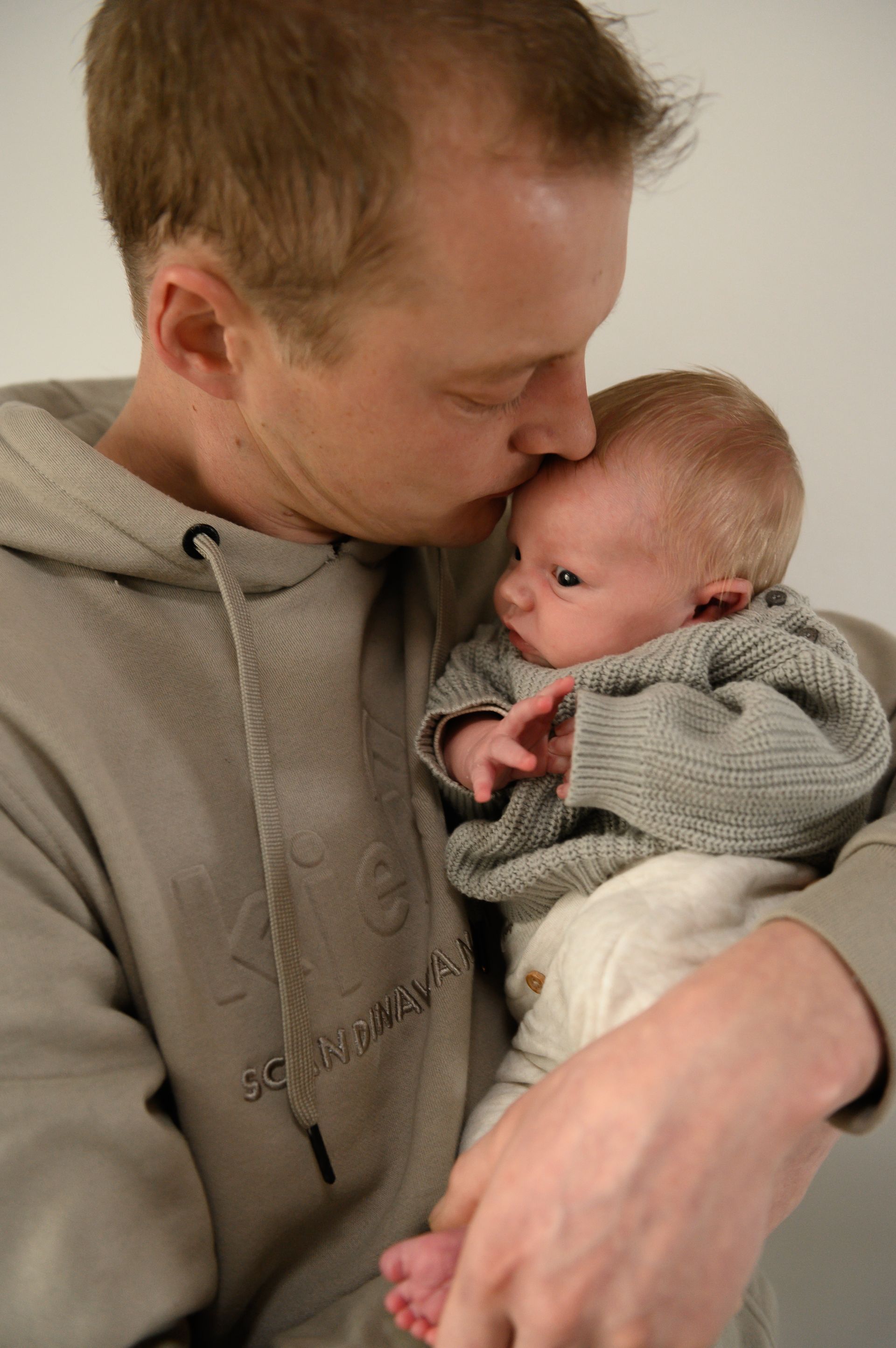 Een man kust een baby op het voorhoofd; beiden dragen grijze truien en staan ​​voor een witte muur.