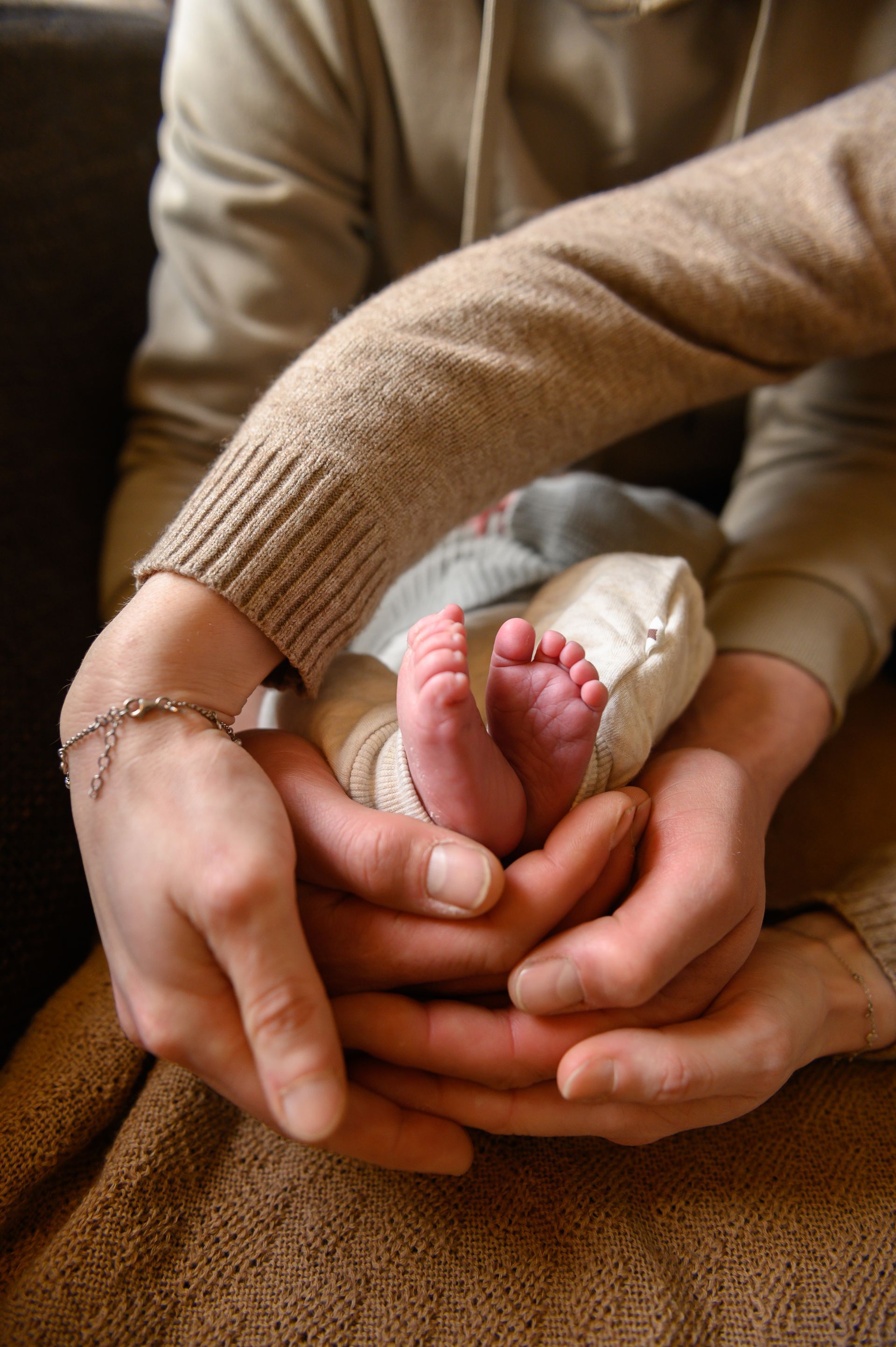 Handen omarmen de kleine voetjes van een baby. De persoon draagt ​​een bruine trui, heeft een zachte huid en een zilveren armband.