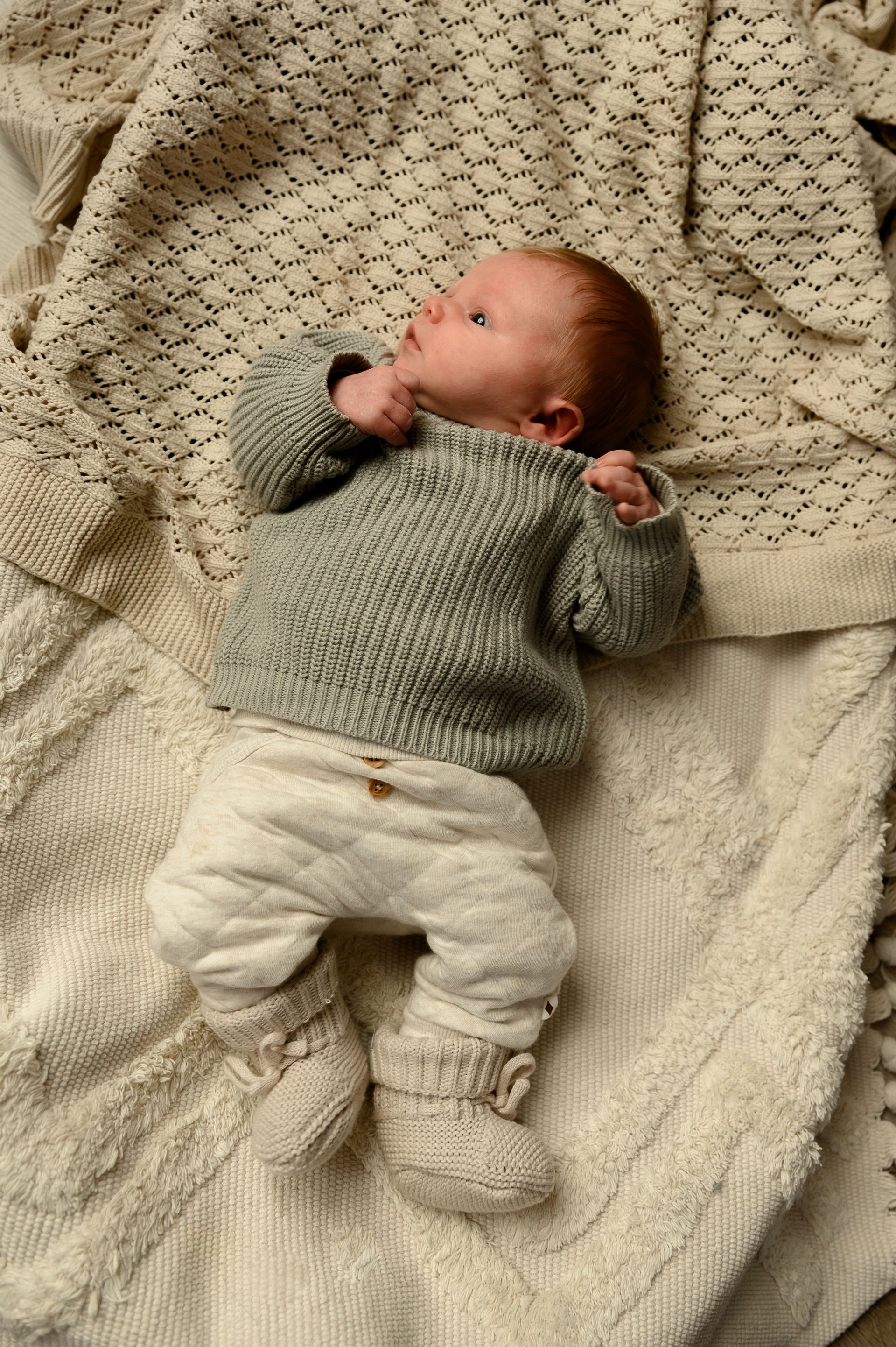 Pasgeboren baby in grijze trui en broek, liggend op een crèmekleurige deken, kijkt omhoog.