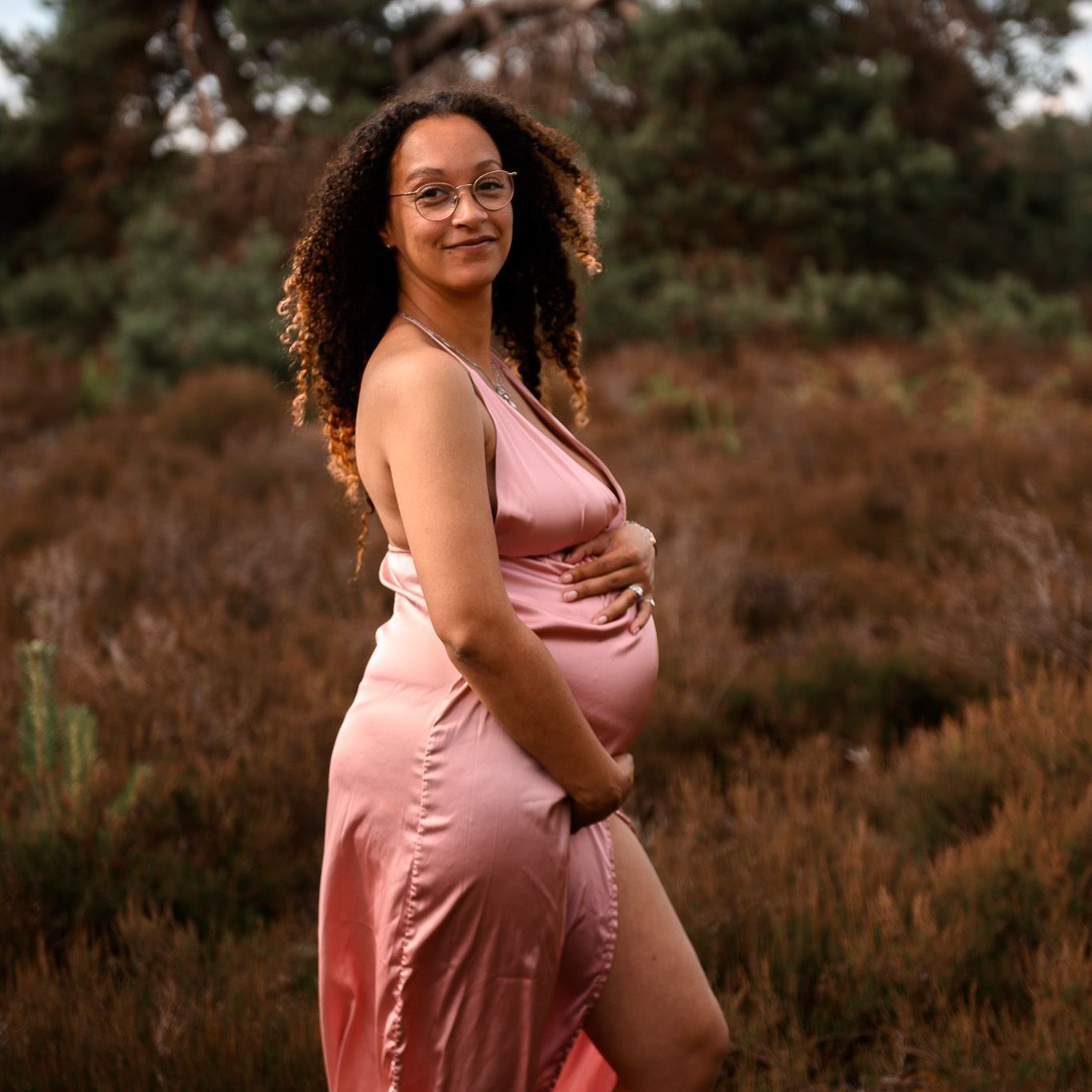 Een zwangere vrouw in een roze satijnen jurk houdt haar buik vast en lacht in een veld met bruine planten.