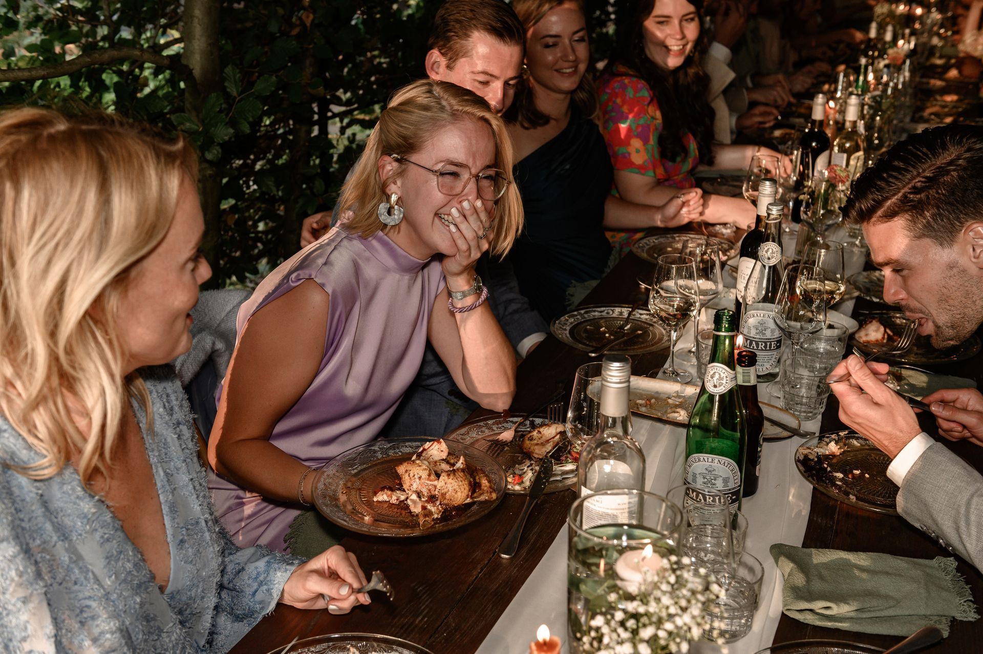 Mensen zitten aan een lange tafel, lachen en eten. Glazen en flessen staan ​​op tafel.