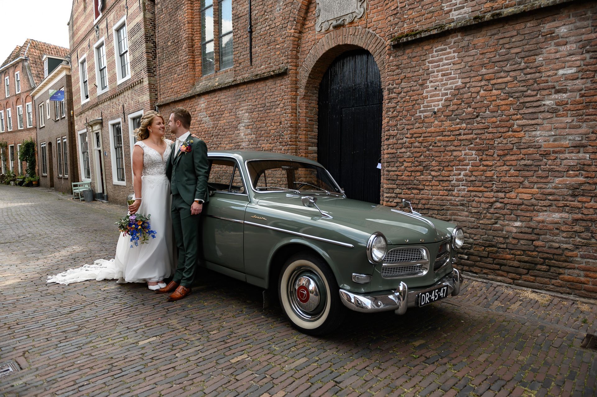Bruid en bruidegom poseren naast een vintage groene auto op een straat met kinderkopjes. Op de achtergrond zijn bakstenen gebouwen te zien.