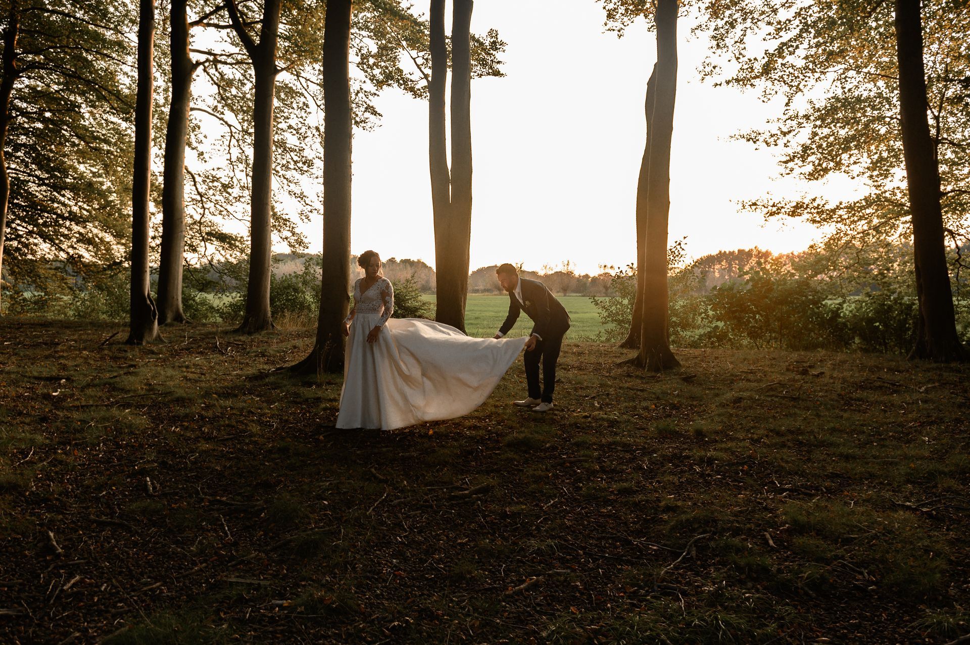 Een bruidspaar in trouwkleding in een bos bij zonsondergang; de bruidegom past de jurk van de bruid aan.