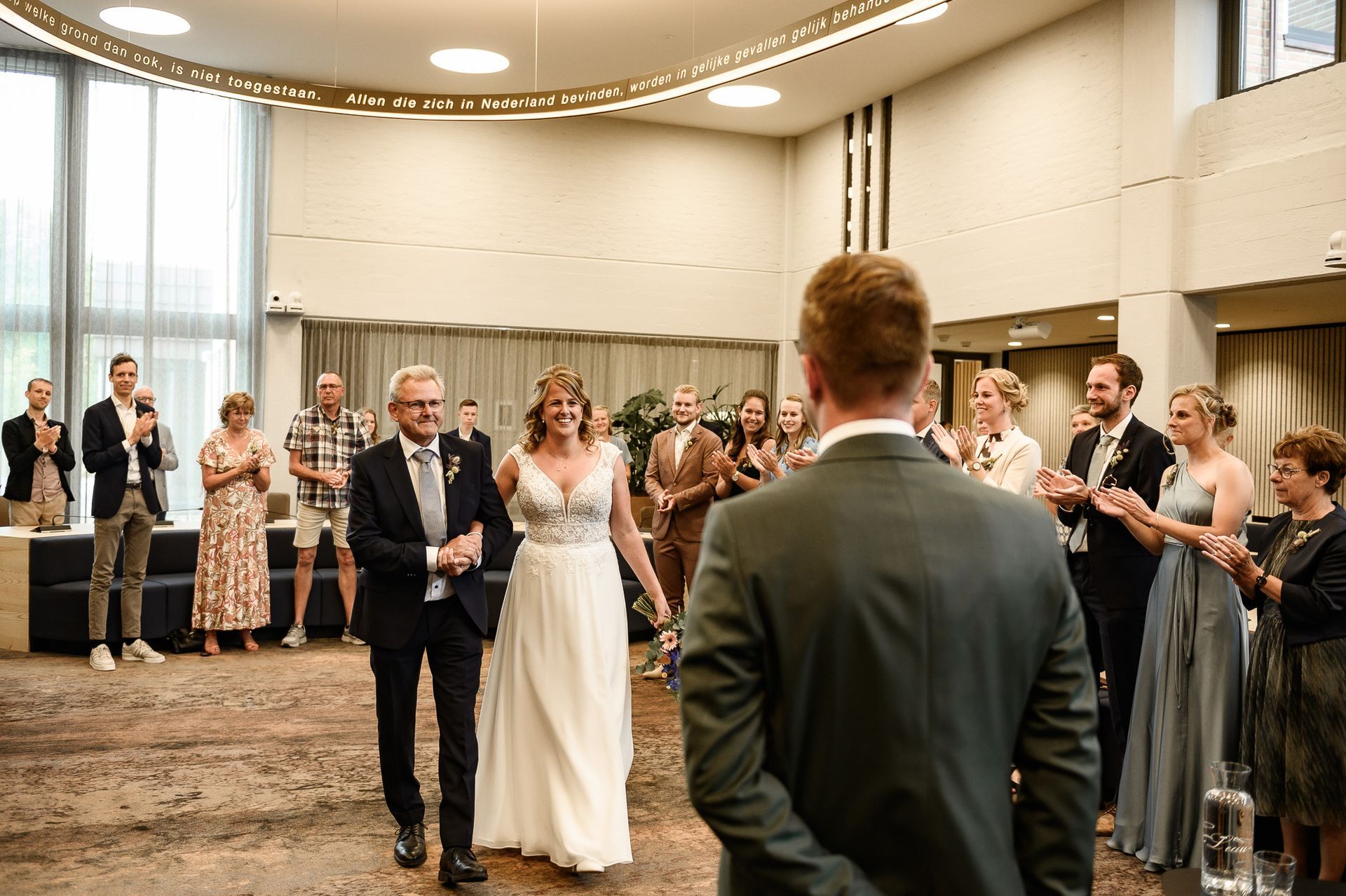 De bruid loopt met haar vader naar de bruidegom toe, de gasten applaudisseren, interieur.