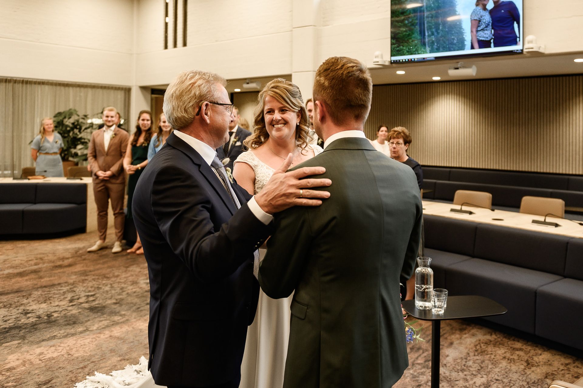 Huwelijksceremonie. Een man in pak legt zijn hand op de schouder van de bruidegom. De bruid glimlacht. Gasten op de achtergrond.