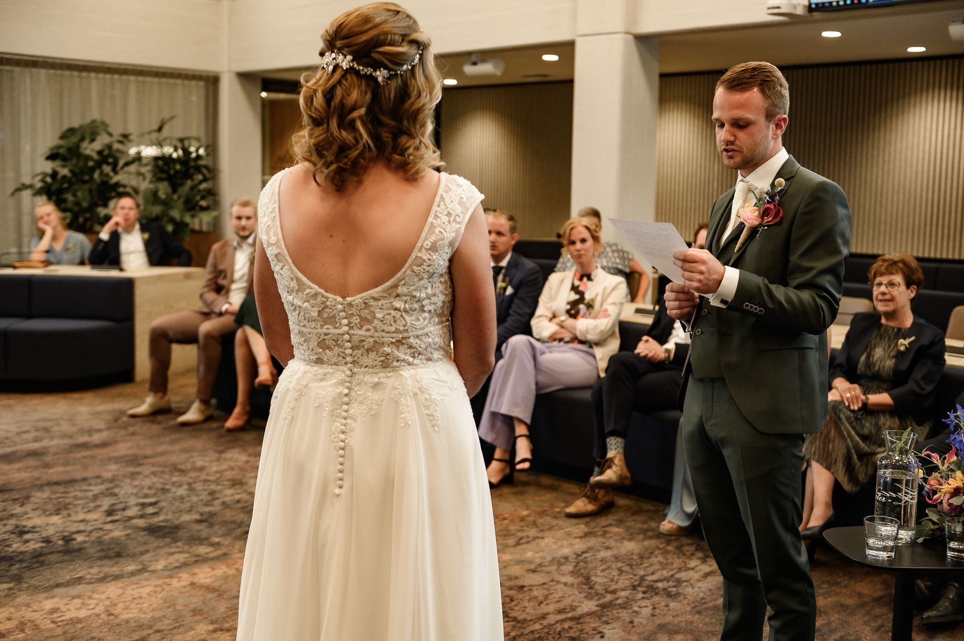 Een man leest zijn huwelijksgeloften voor aan een vrouw tijdens een huwelijksceremonie. Gasten zitten; het is een binnenlocatie.